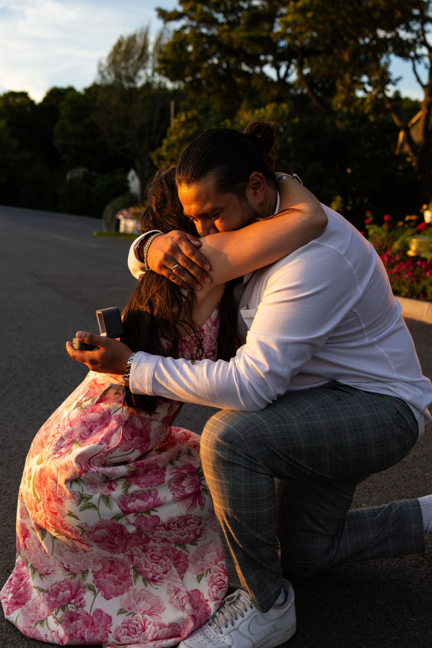 Man on one knee hugging woman during outdoor marriage proposal at golden hour, holding ring box.