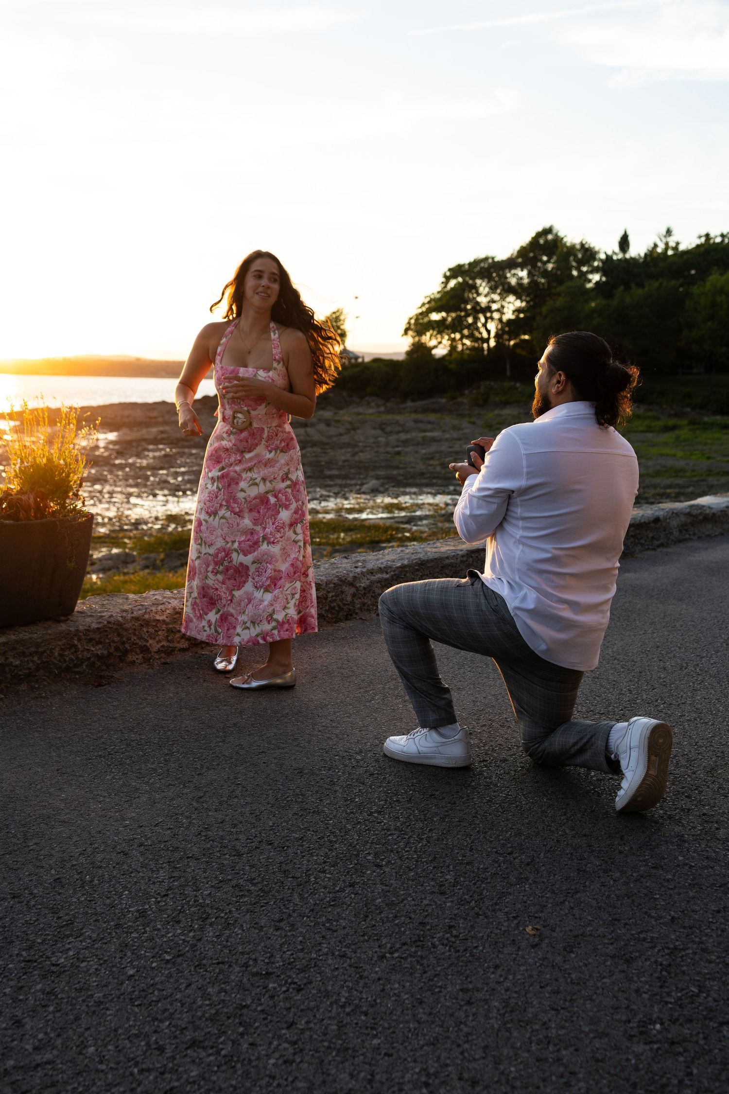 Man proposes on one knee to woman in floral dress at sunset by the ocean, golden light glowing behind them.