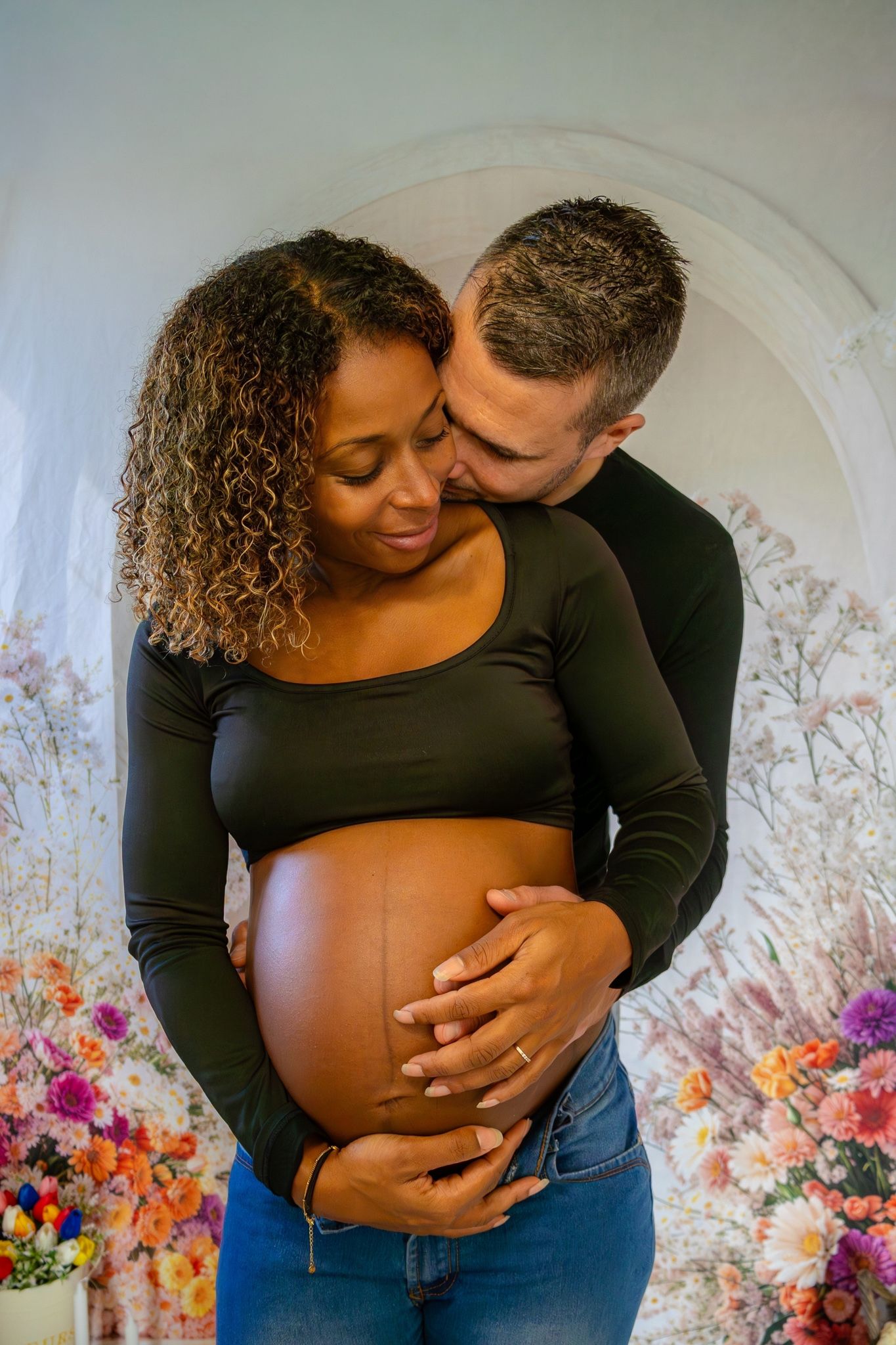 Man tenderly kissing pregnant woman's head while embracing her baby bump before colorful floral backdrop.