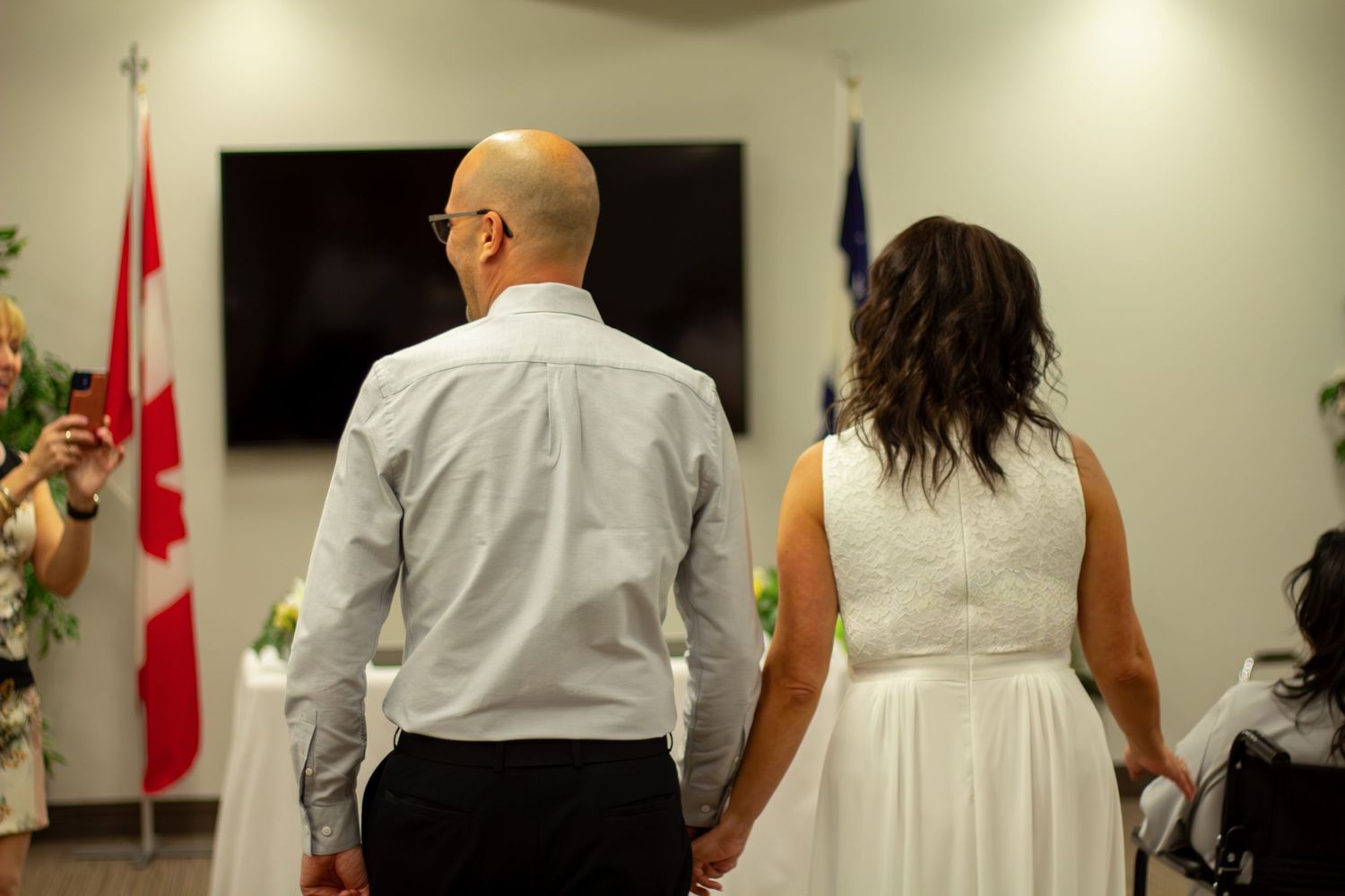 Newlywed couple holds hands facing officiant with Canadian flag during indoor ceremony.