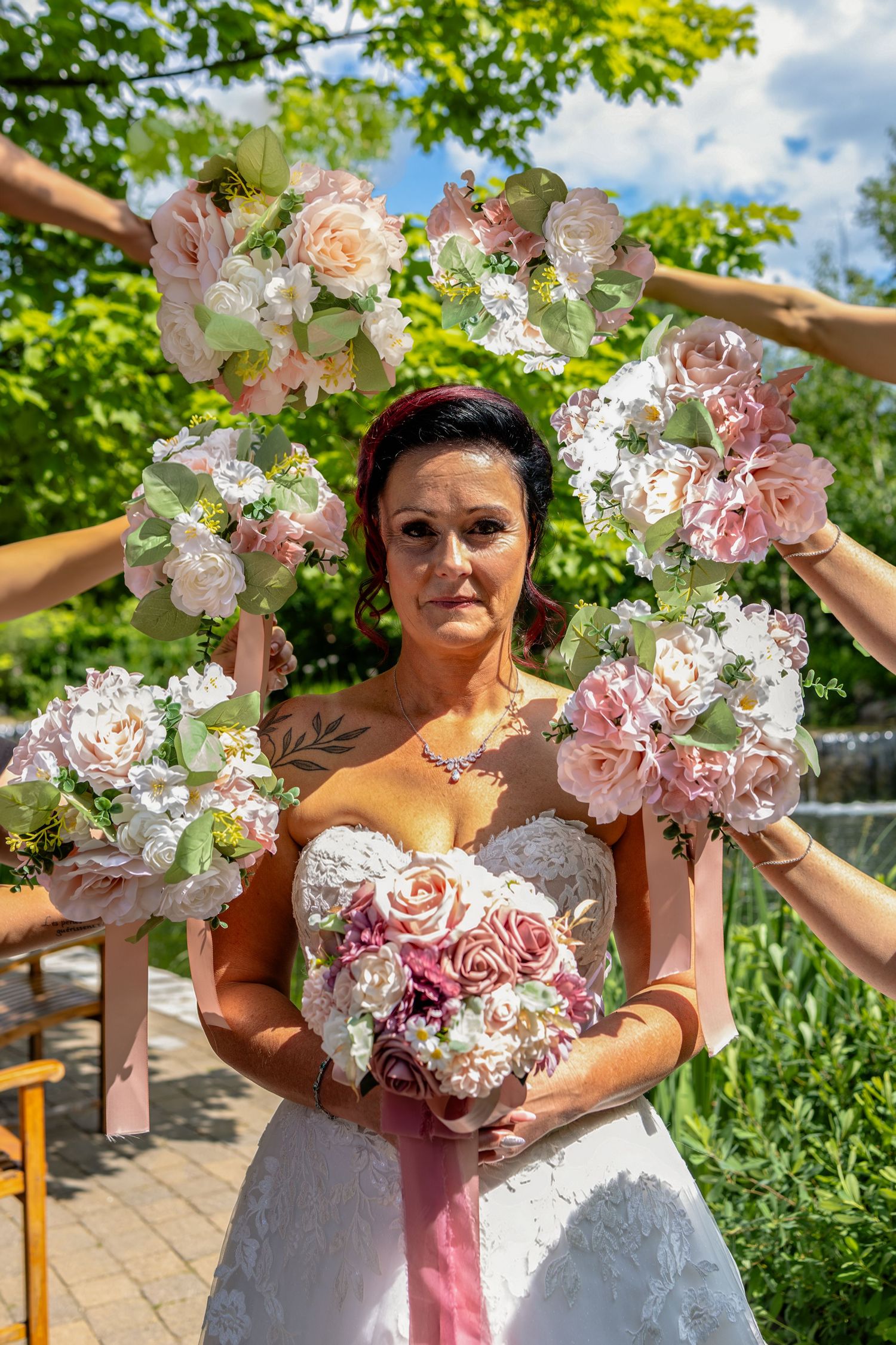 Bride in white gown holds pink bouquet, framed by bridesmaids holding floral wreaths outdoors.