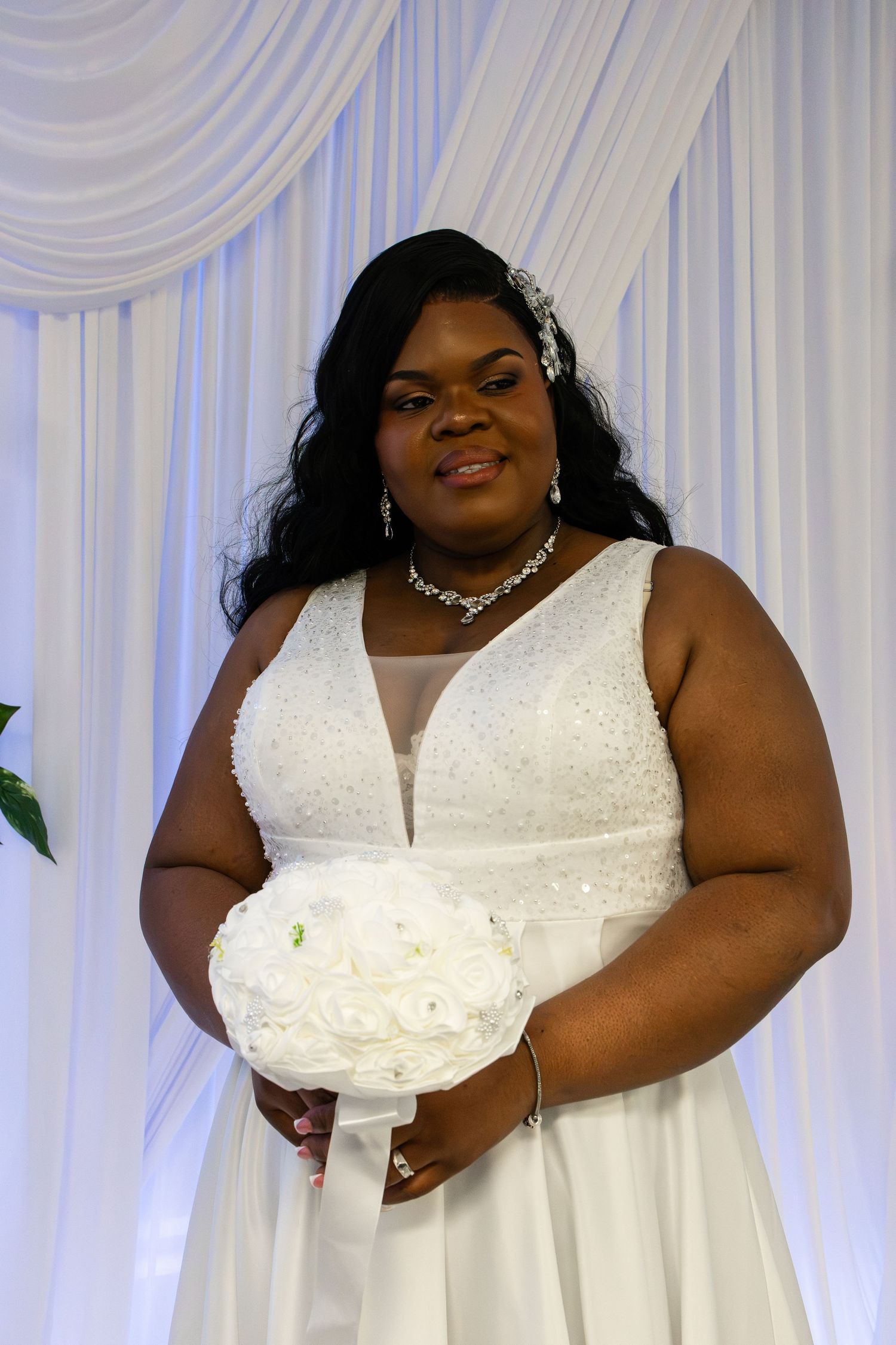 A bride in a white beaded gown holds a white bouquet, wearing crystal jewelry against a draped white backdrop.