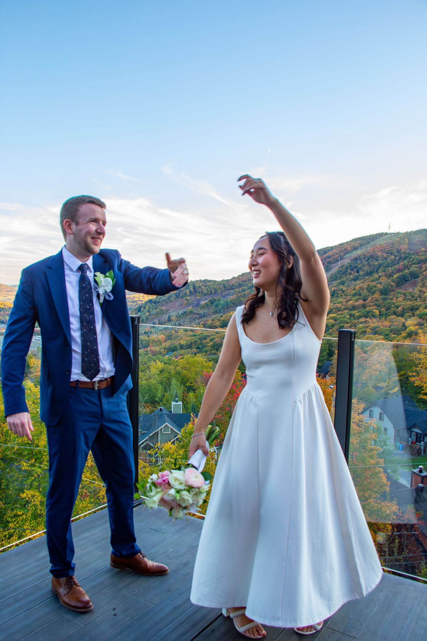 Joyful bride and groom celebrate on a deck with stunning fall mountain scenery, raising glasses and bouquet.