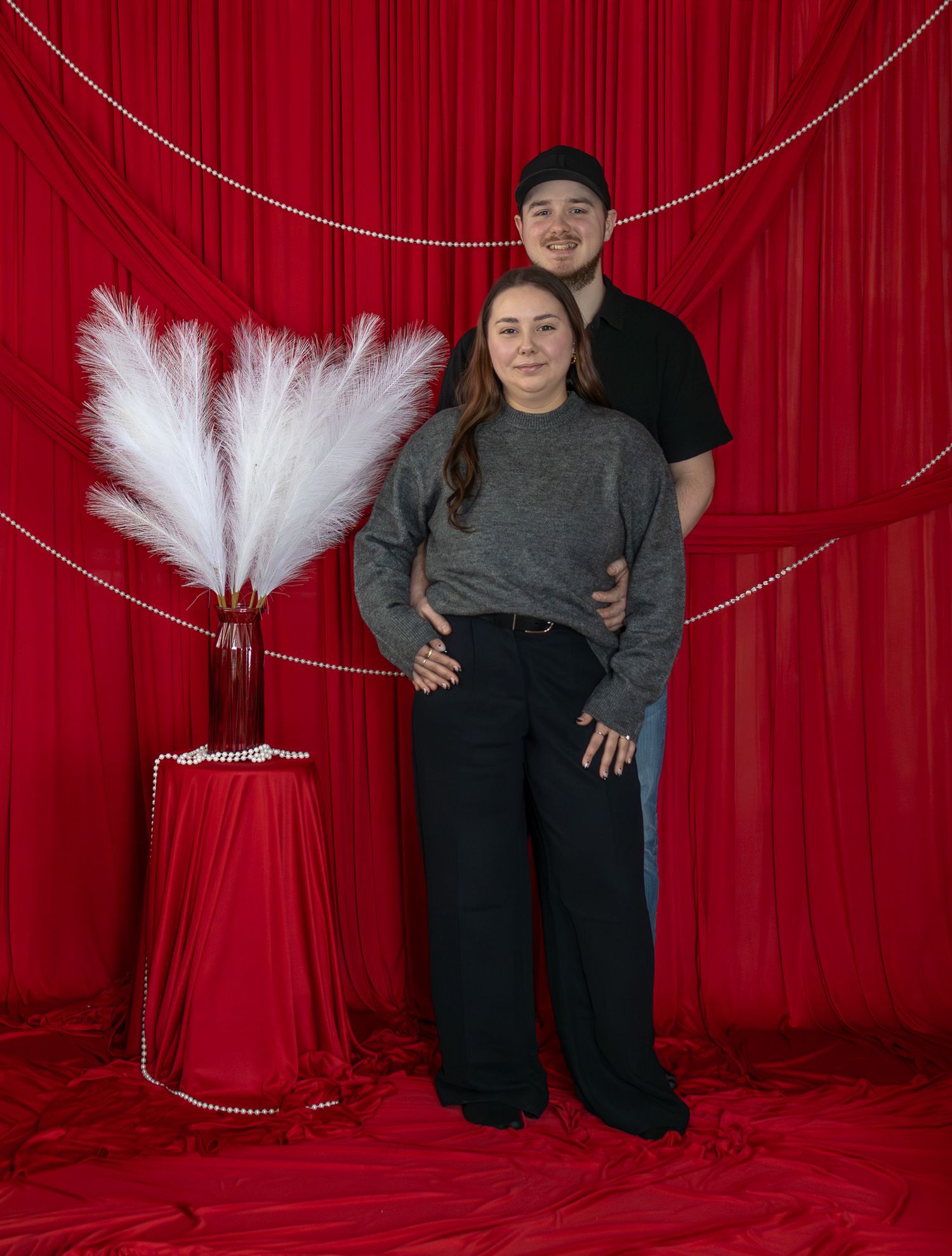 Couple posing before red curtain backdrop with white pampas grass and pearl garland decor.
