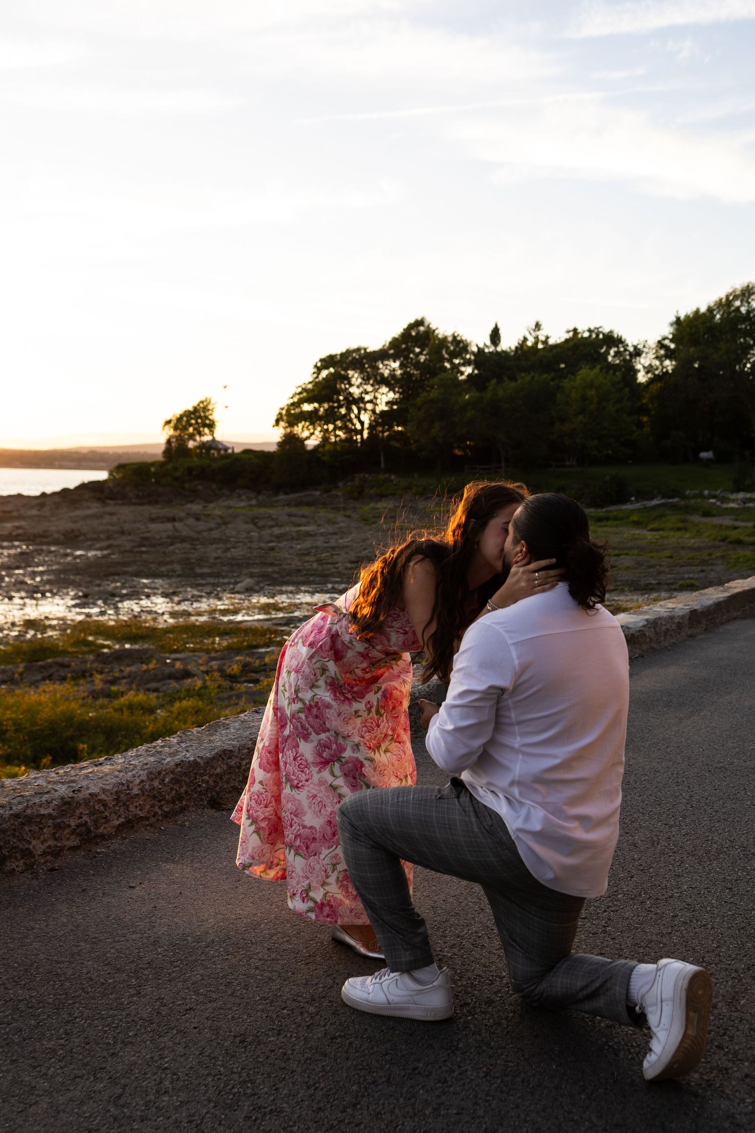 Couple sharing a romantic kiss at sunset by the waterfront, man kneeling in white shirt and gray pants.