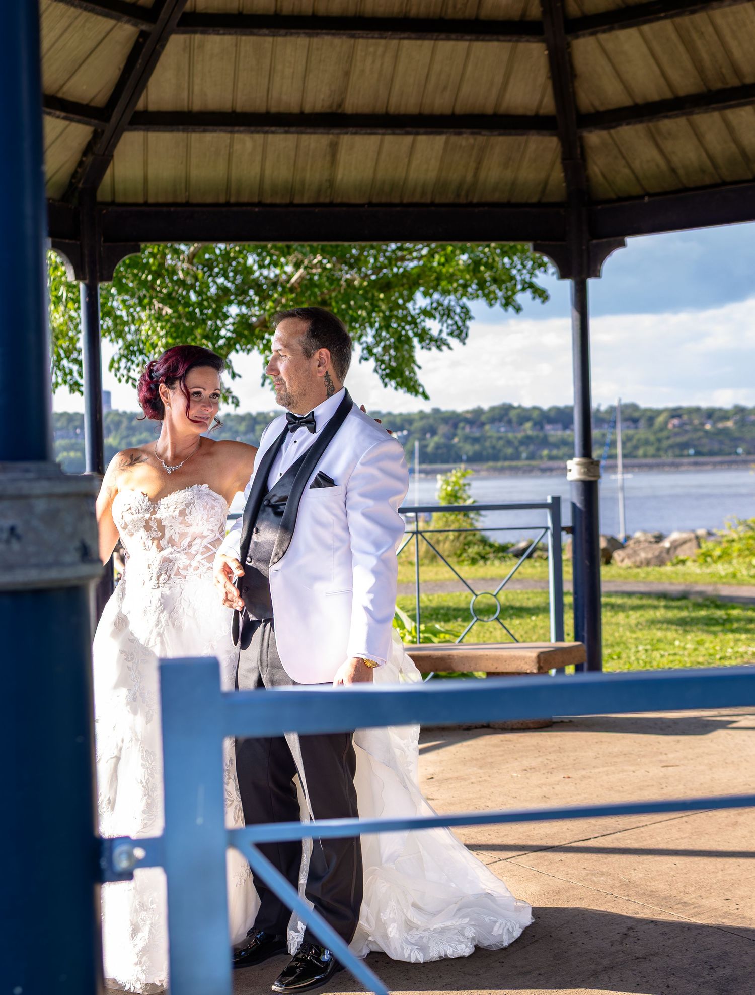 Bride and groom pose under a waterfront gazebo, she in a lace gown, he in a white tuxedo jacket.