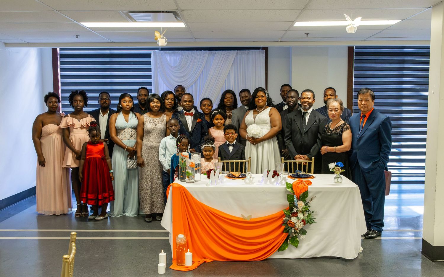 Large wedding group photo with guests posed around a decorated table with orange and white tablecloth.