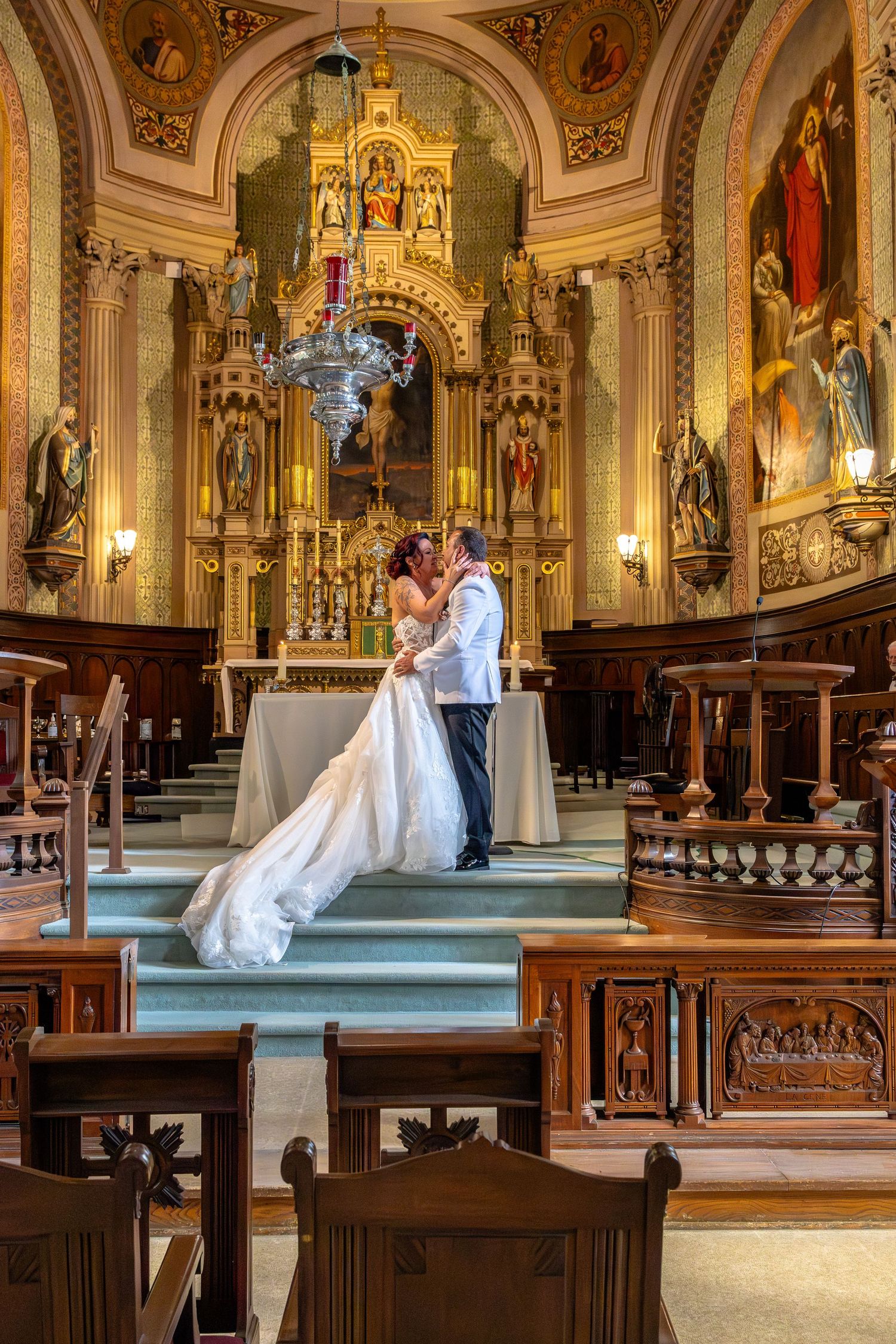 Bride and groom share a kiss at the altar of an ornate church with golden decor and religious artwork.