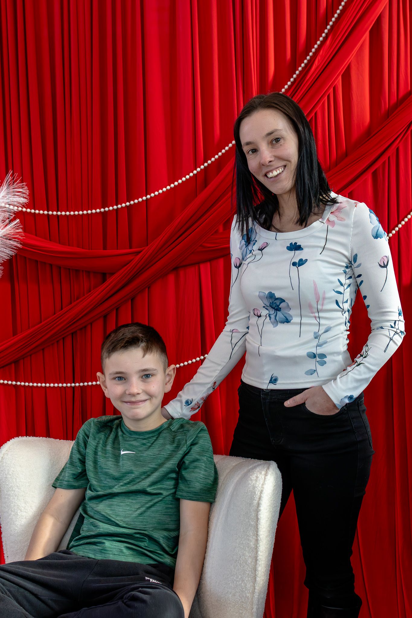 Smiling woman in floral top standing beside young boy seated in chair against red curtains.