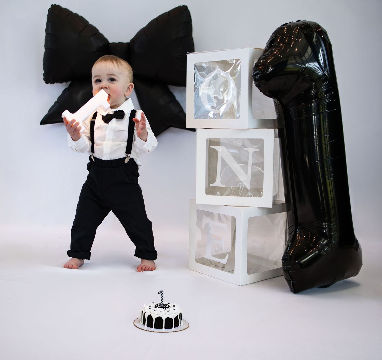 Baby boy in suspenders celebrates first birthday with letter N boxes, number 1 balloon, and smash cake.