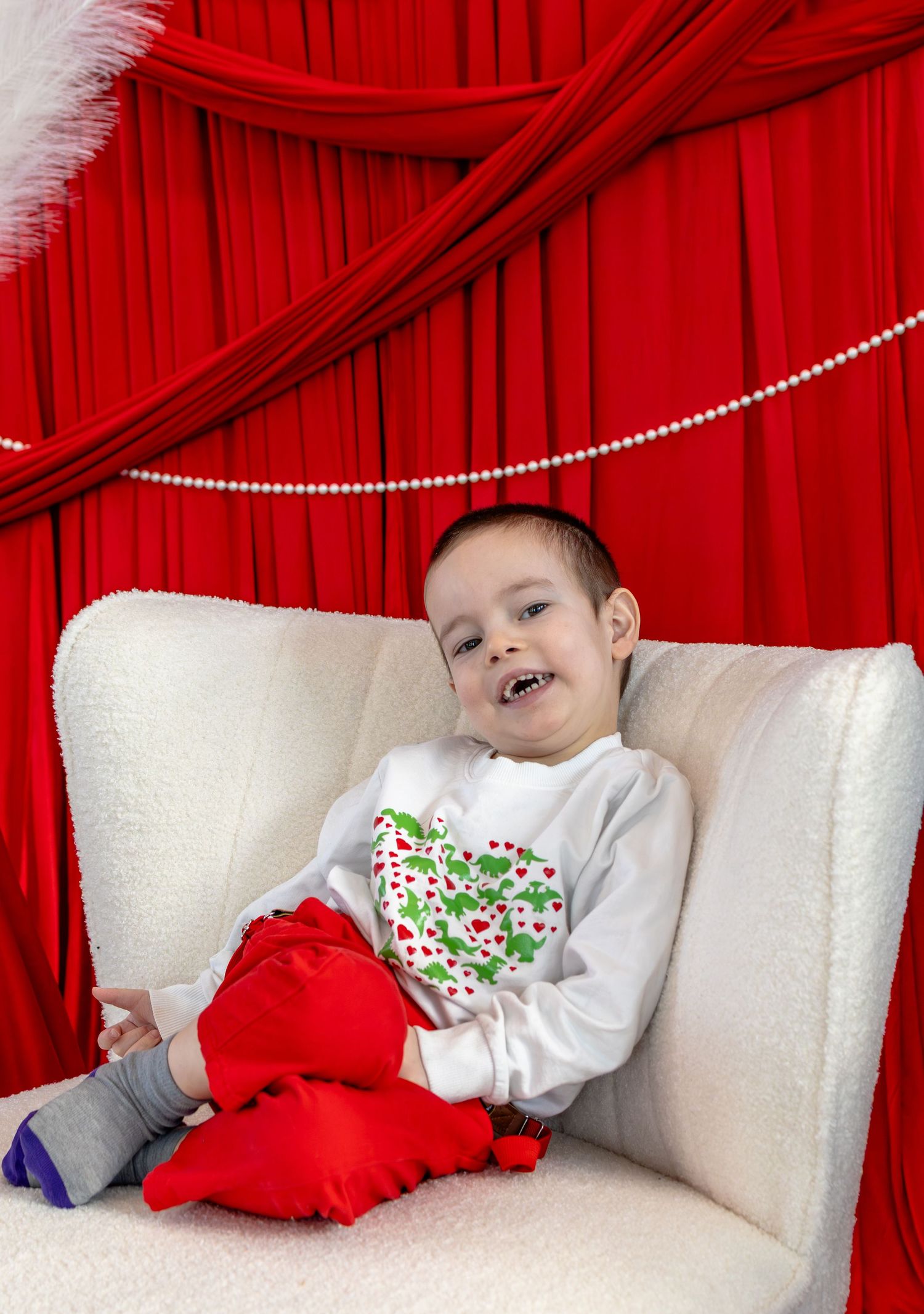 A cheerful young boy sits smiling in a white chair with red holiday decor and a Christmas-themed shirt.