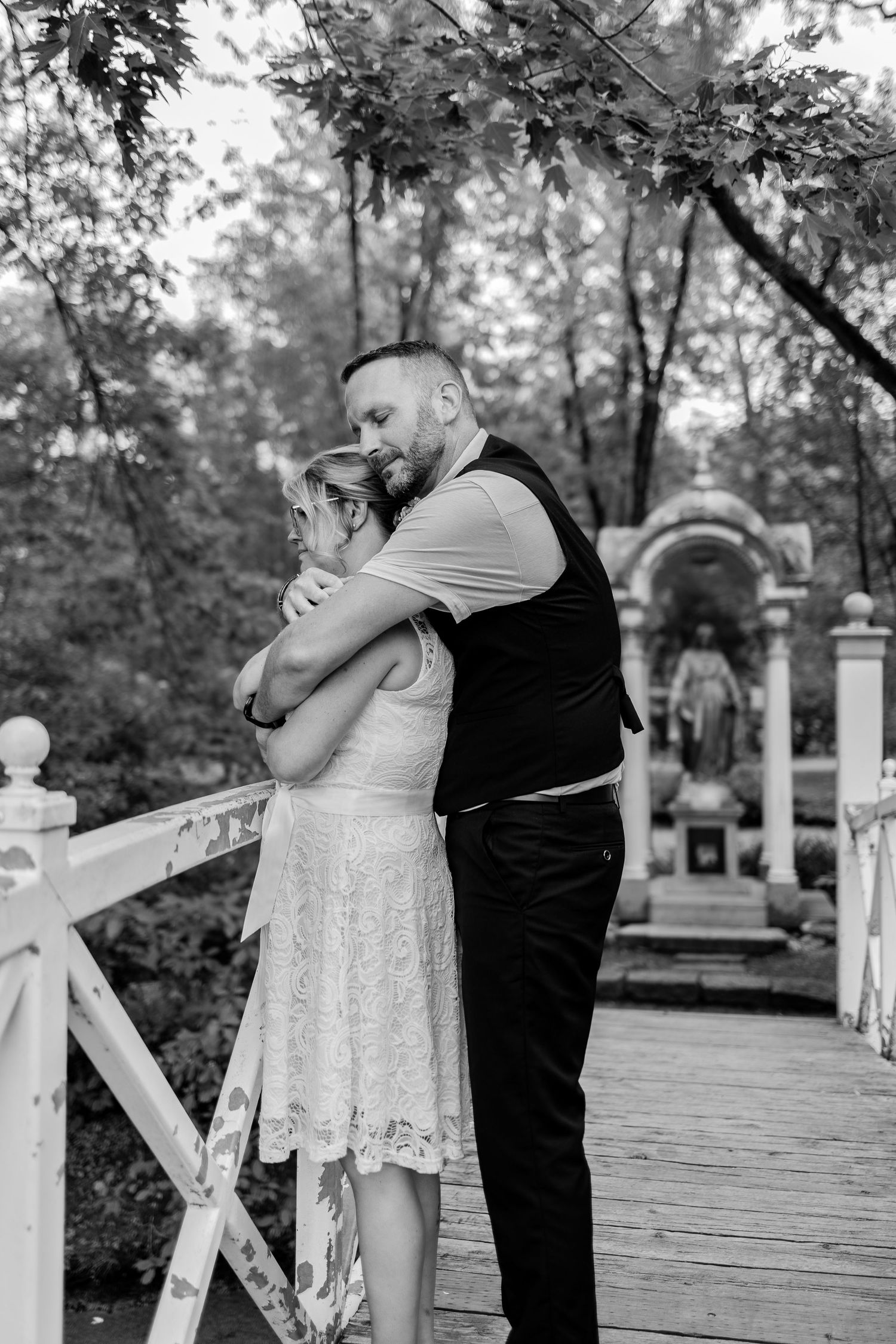 Black and white photo of a couple embracing on a wooden bridge in a lush garden near a decorative statue.