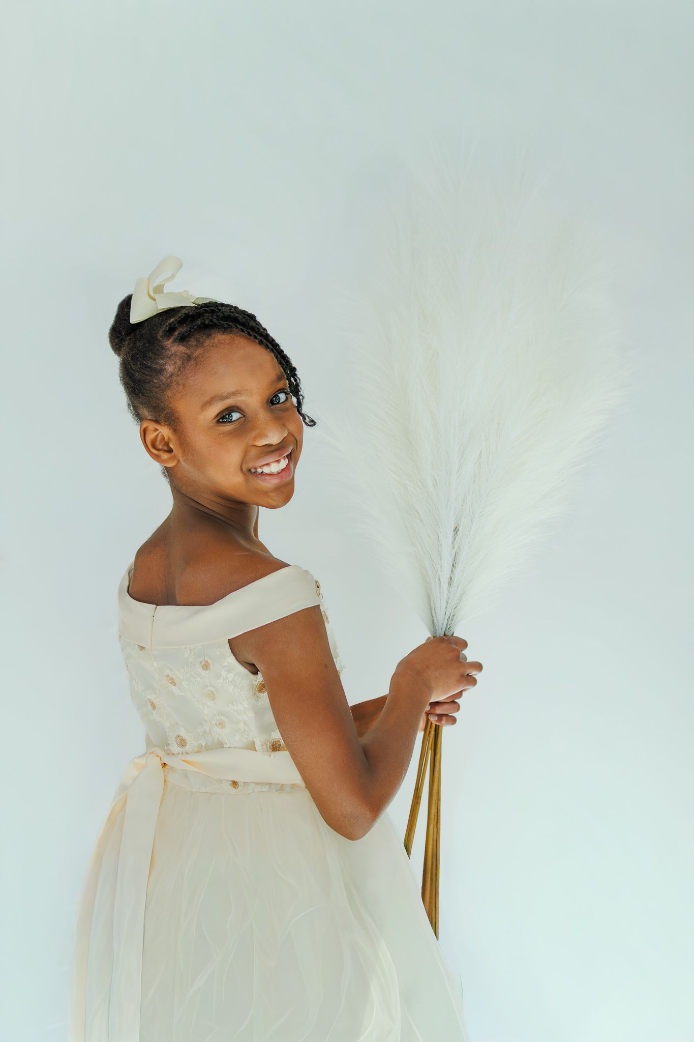 Smiling young Black girl in white dress holding white pampas grass bouquet with gold ribbon.