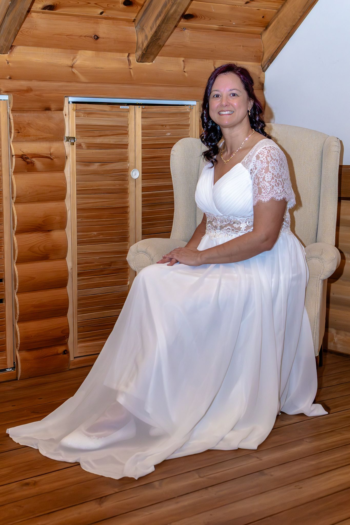 Smiling bride in a white lace wedding gown sitting gracefully in a cream armchair inside a log cabin.