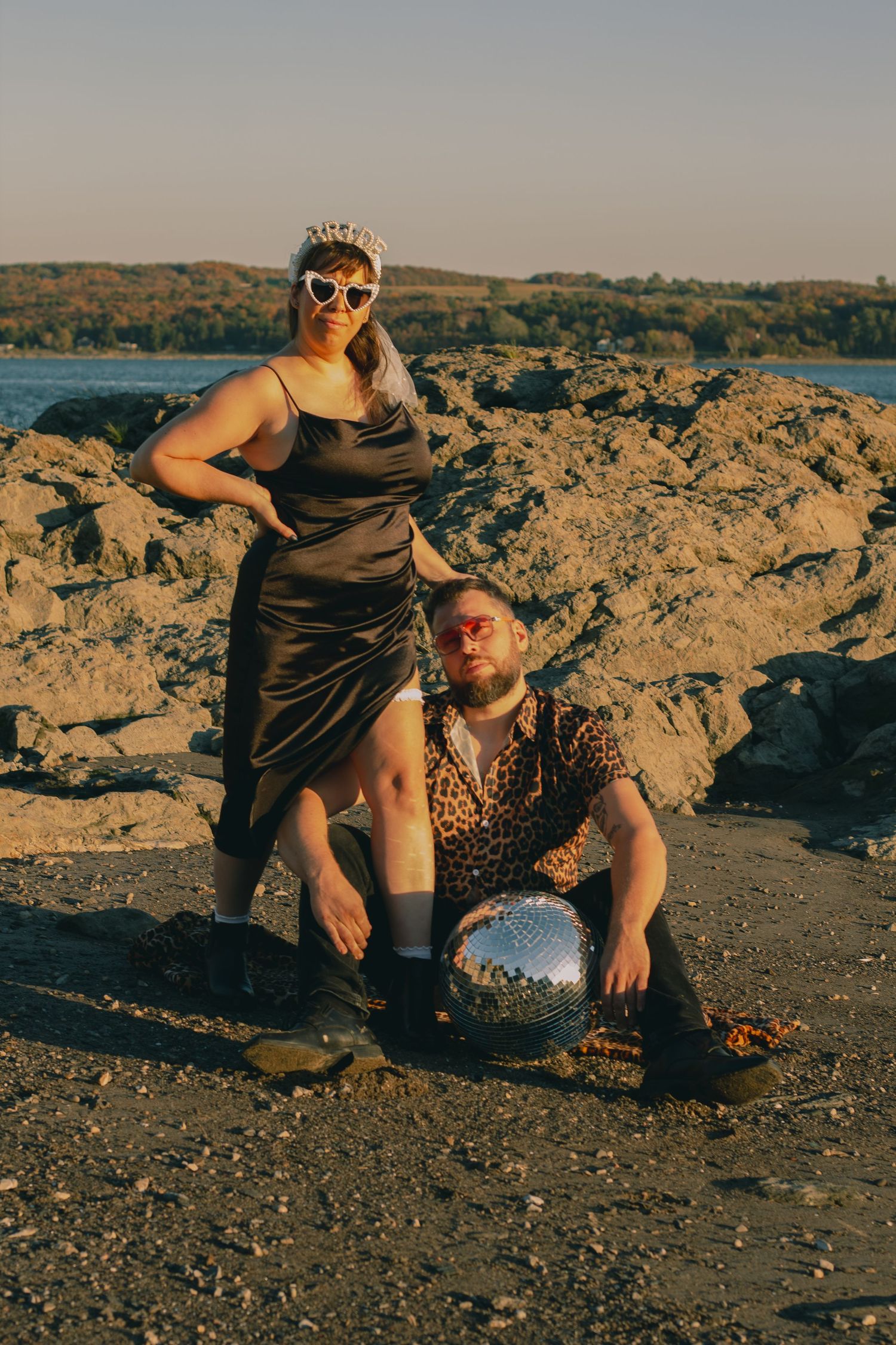 A couple poses on a rocky beach, woman in silver dress with tiara, man in leopard shirt holding a disco ball.