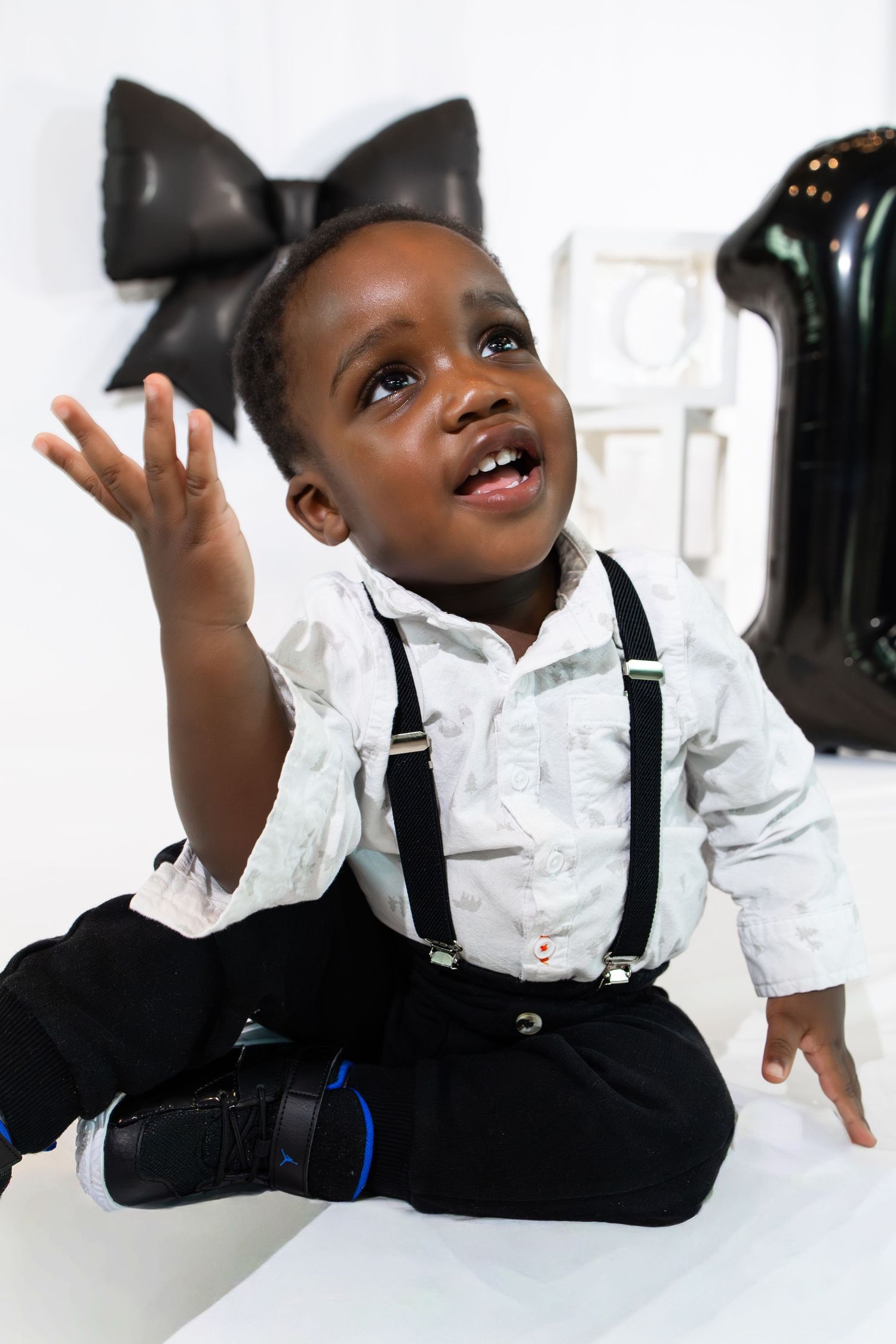 A joyful Black toddler boy in suspenders and white shirt reaches up during his first birthday photo shoot.