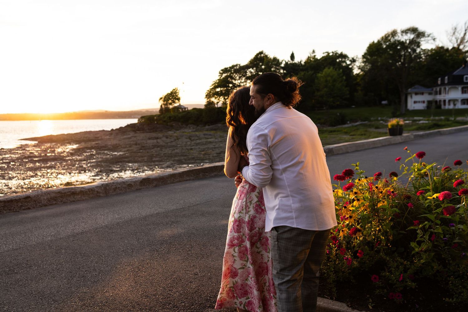 Couple embracing at sunset by the waterfront with colorful flowers and golden light in the background.