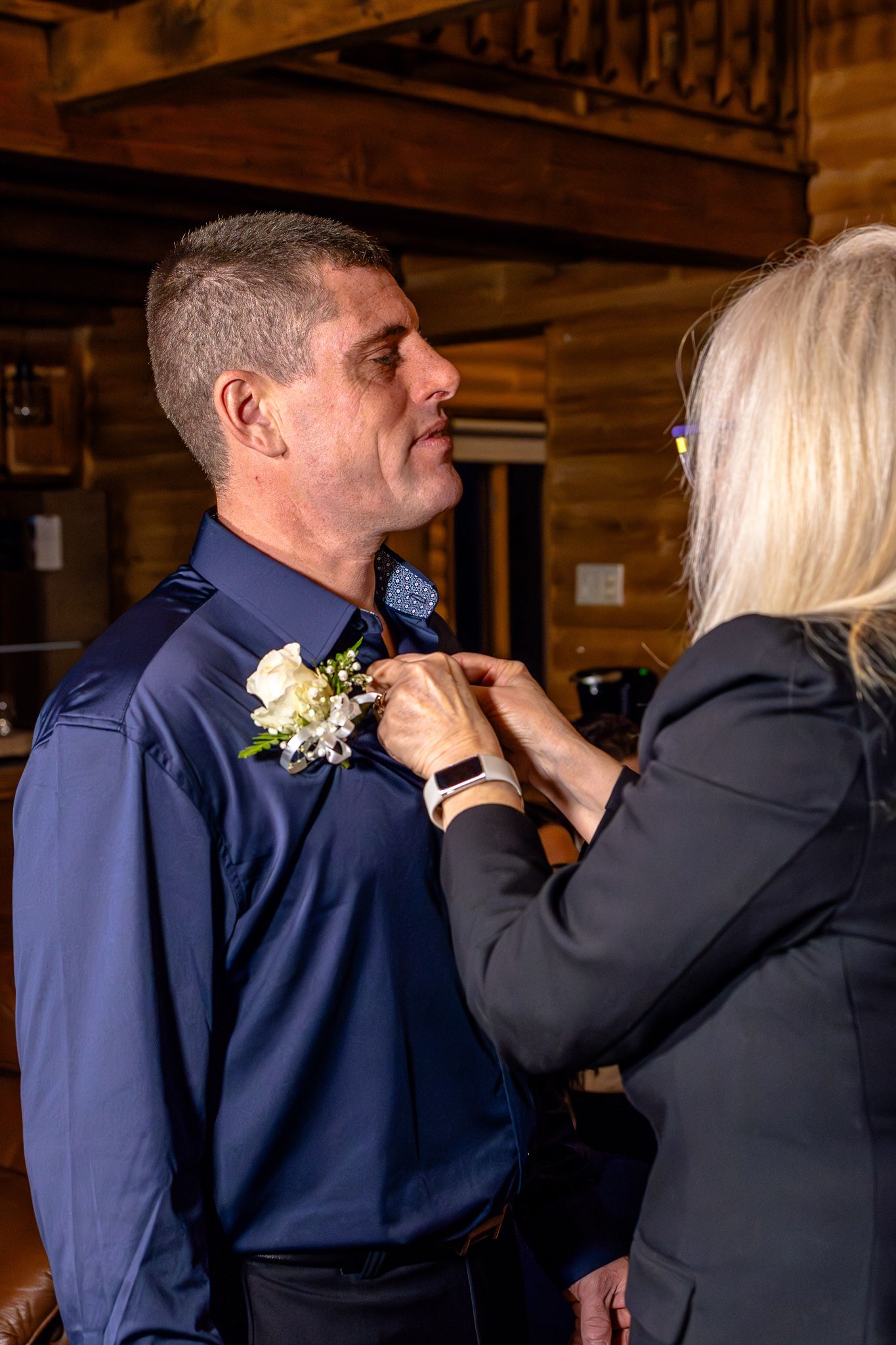 Blonde woman pinning a white rose boutonniere on a man in a navy blue dress shirt at a wedding.