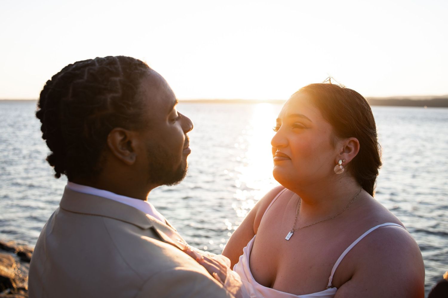 Couple sharing a romantic moment at sunset by the water, dressed in wedding attire, golden light glowing behind them.