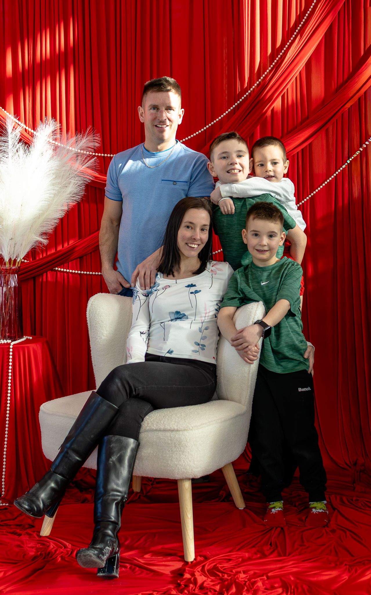 Happy family of five posing together on a white chair with elegant red curtain backdrop.