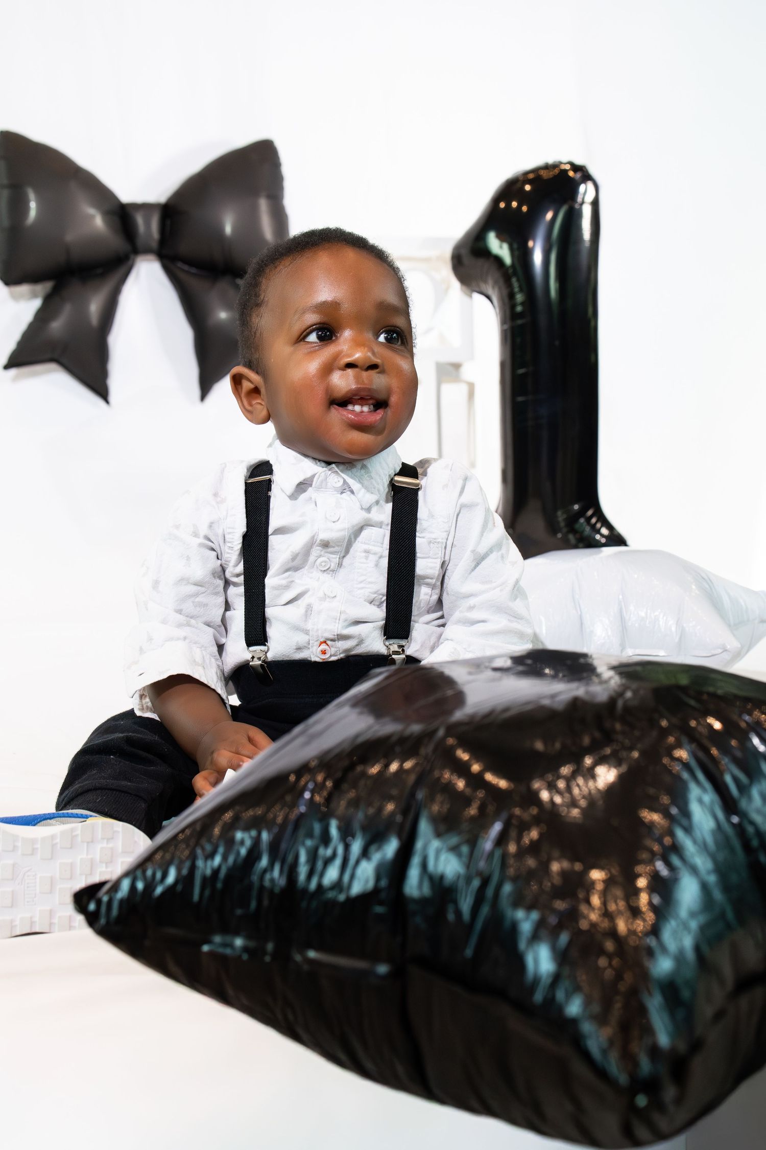 Adorable baby boy in suspenders sits by a black number 1 balloon celebrating his first birthday.
