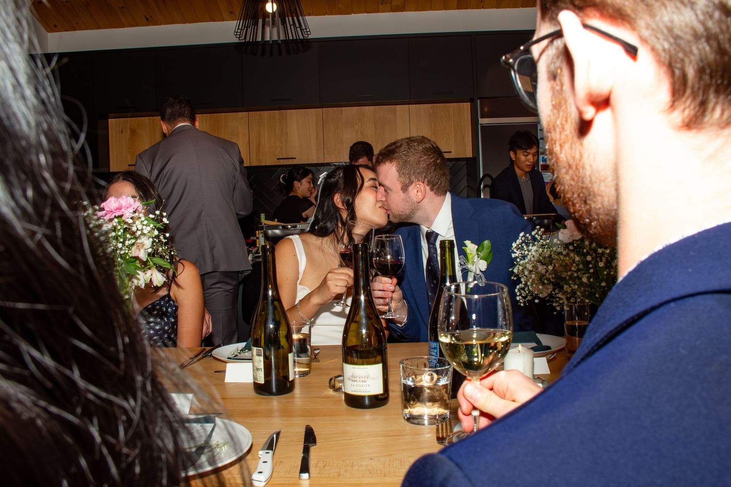 Bride and groom share a kiss at the wedding reception dinner table surrounded by guests and wine.