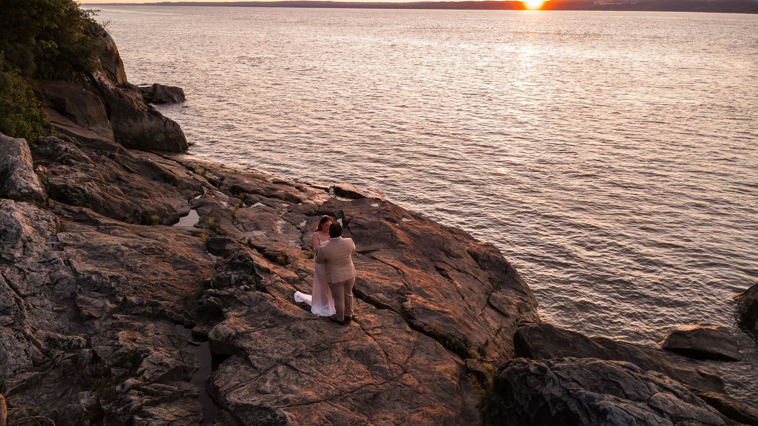 Person standing on rocky shoreline photographing a golden sunset over calm water.