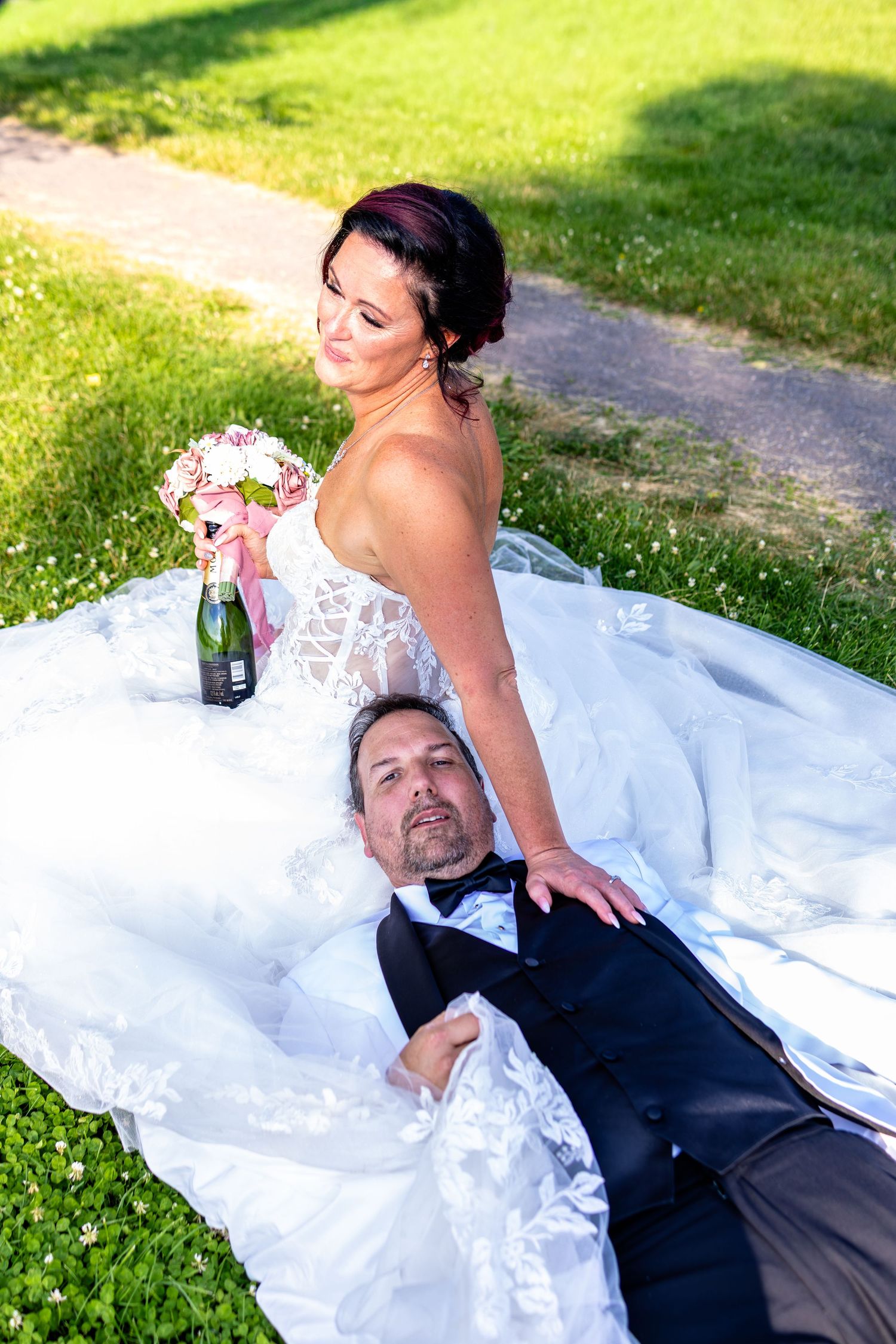 Bride in white lace gown holds bouquet and champagne while groom in tuxedo lies on her dress on green grass.