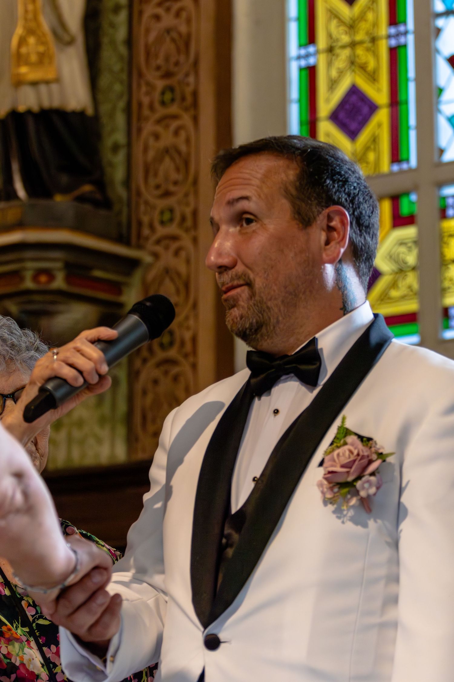Groom in white tuxedo with boutonniere speaks at wedding ceremony in ornate church with stained glass windows.