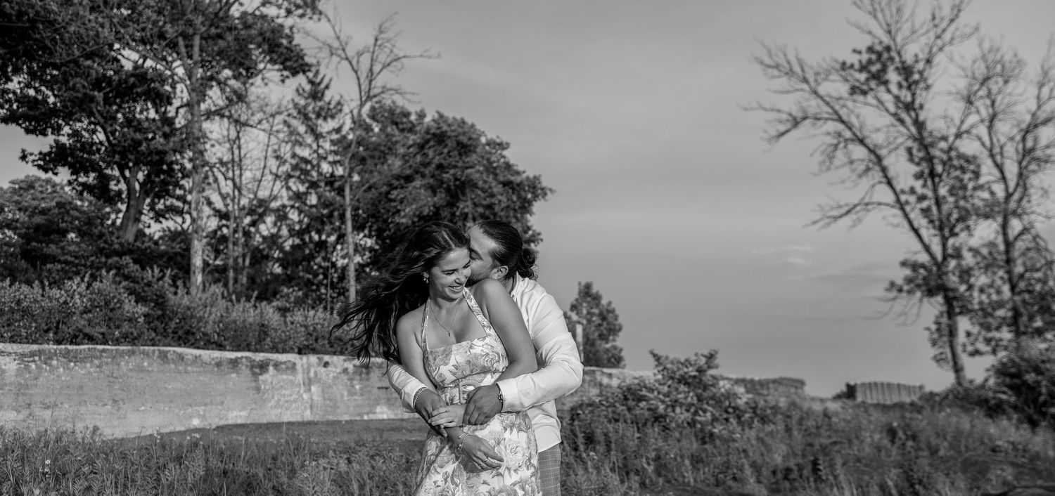 Black and white photo of a couple embracing in a field with trees and cloudy sky in the background.