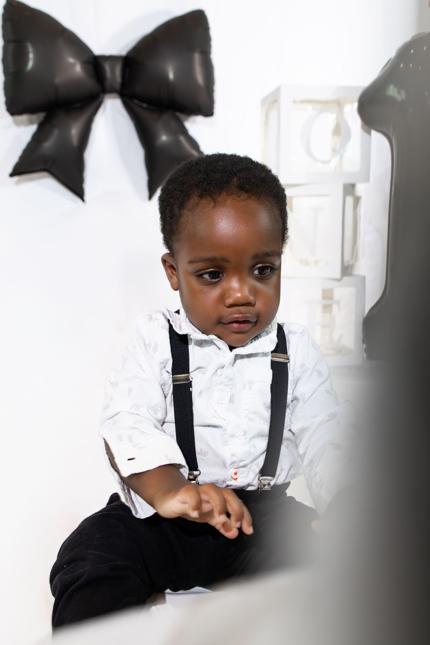 Young Black toddler boy in white shirt with suspenders and black pants sitting during a photo shoot.