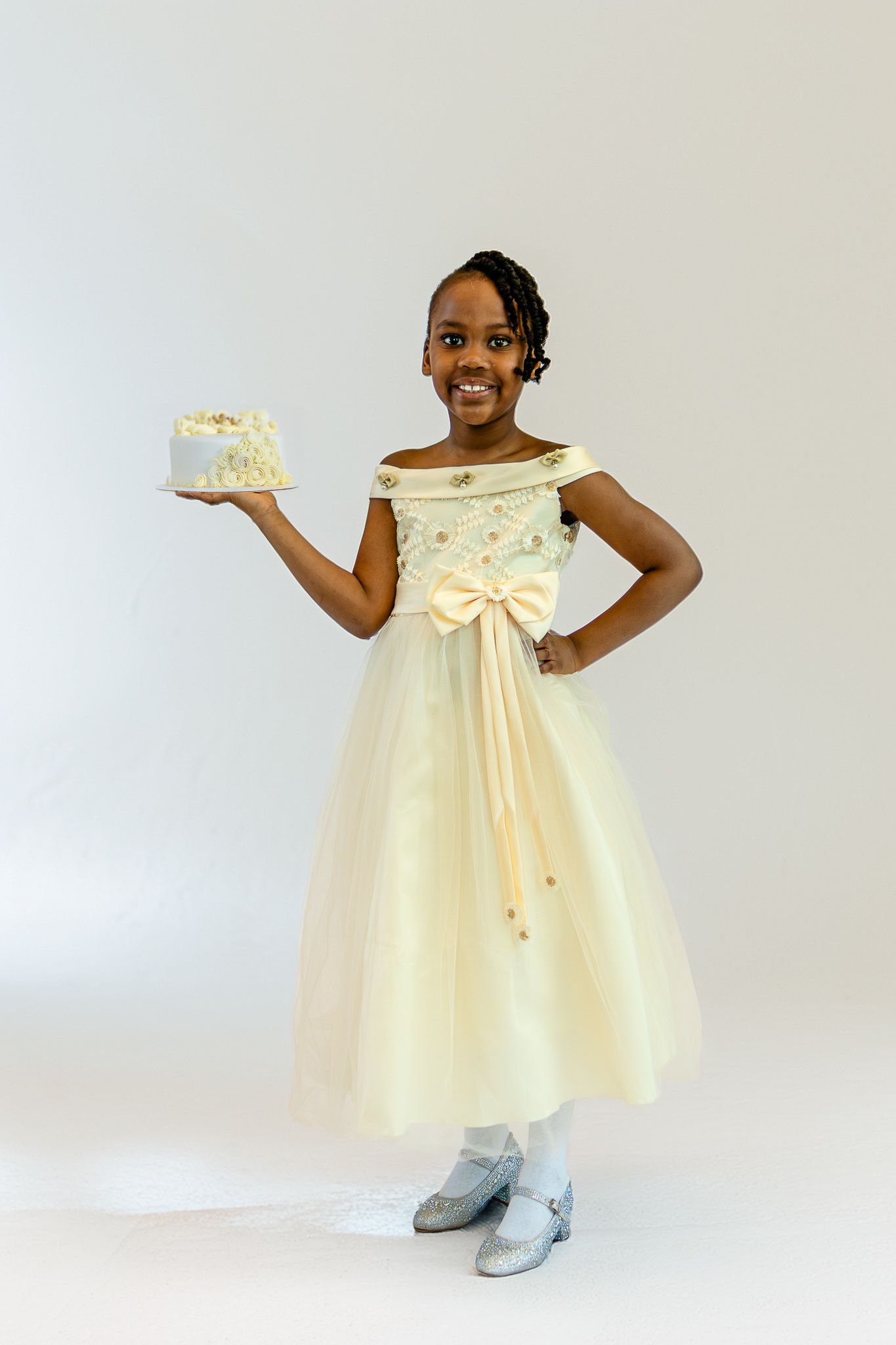 Young Black girl in elegant cream ball gown holding birthday cake, smiling against white background.