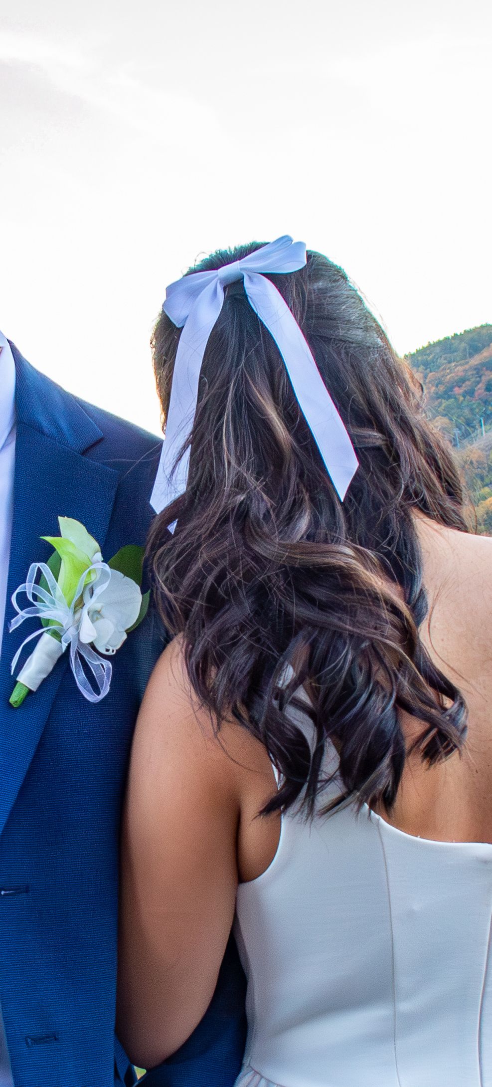 Close-up of brides dark curly hair with white bow and grooms blue suit boutonniere.