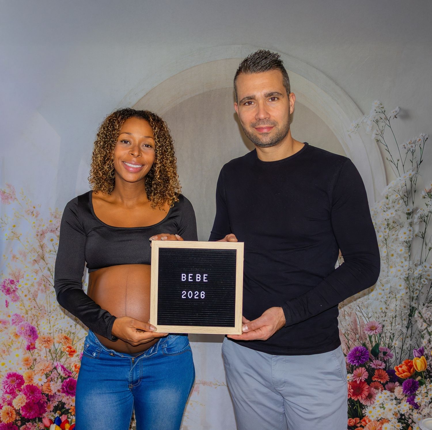 Couple announcing pregnancy with a letter board sign reading BEBE 2026 surrounded by colorful floral backdrop.