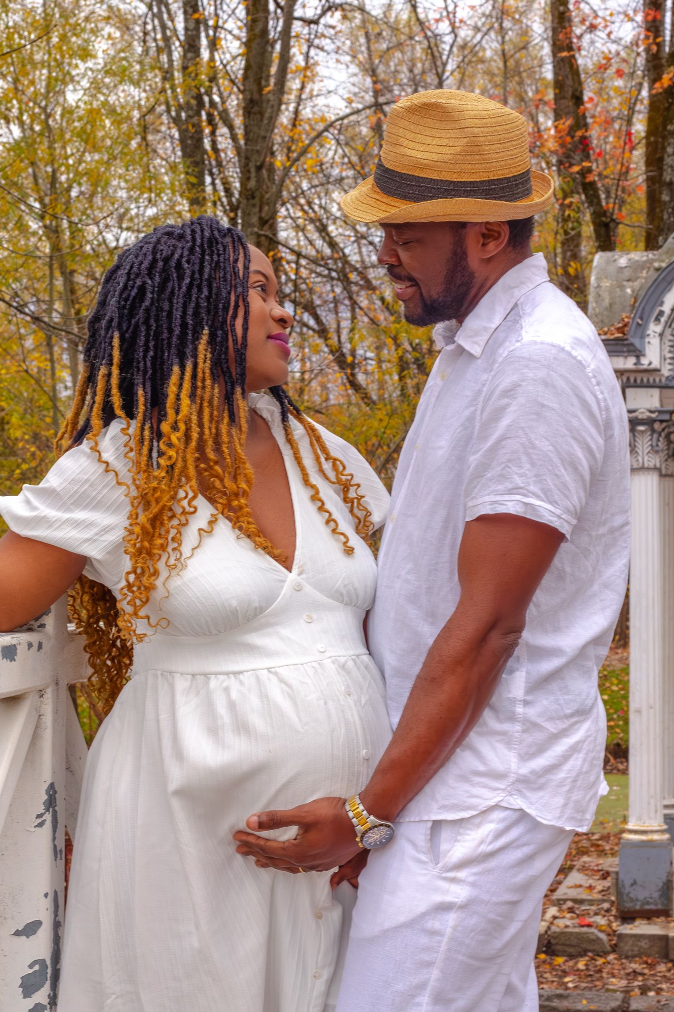 Couple sharing a tender moment together in an autumn park, woman in white maternity dress with colorful braids.