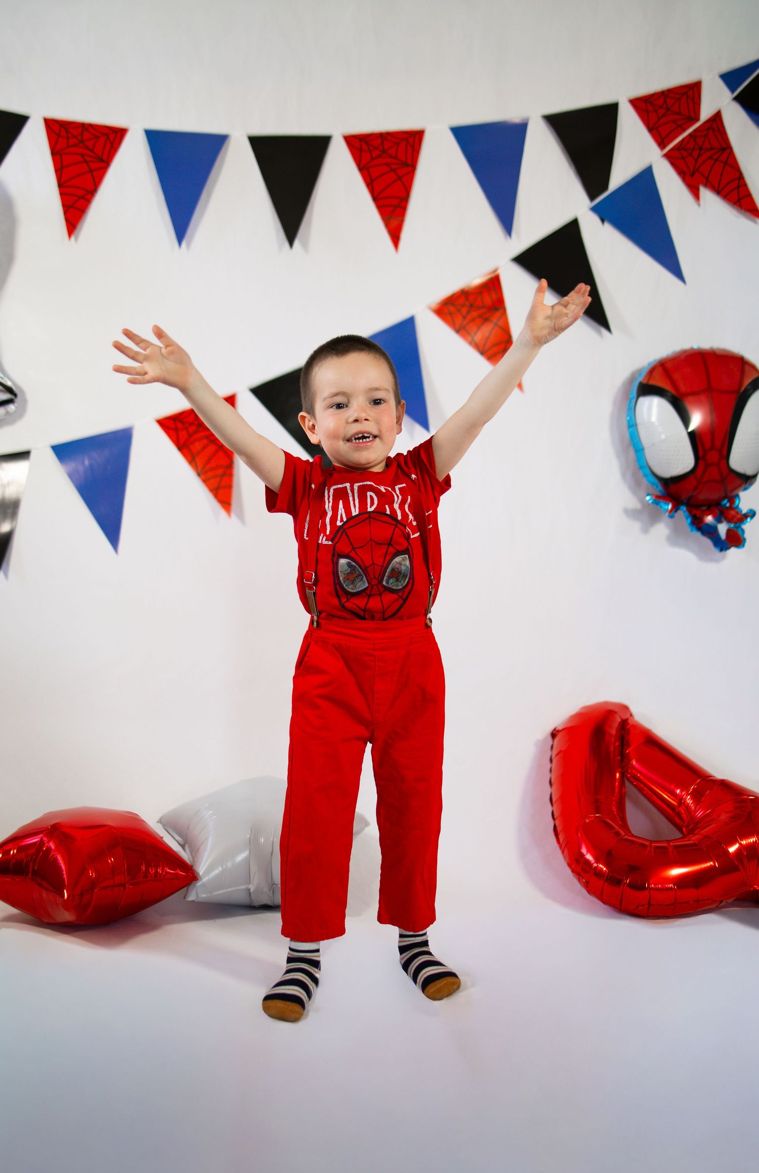 Young boy in red Spider-Man outfit celebrates birthday with colorful bunting and Spiderman balloons, arms raised joyfully.