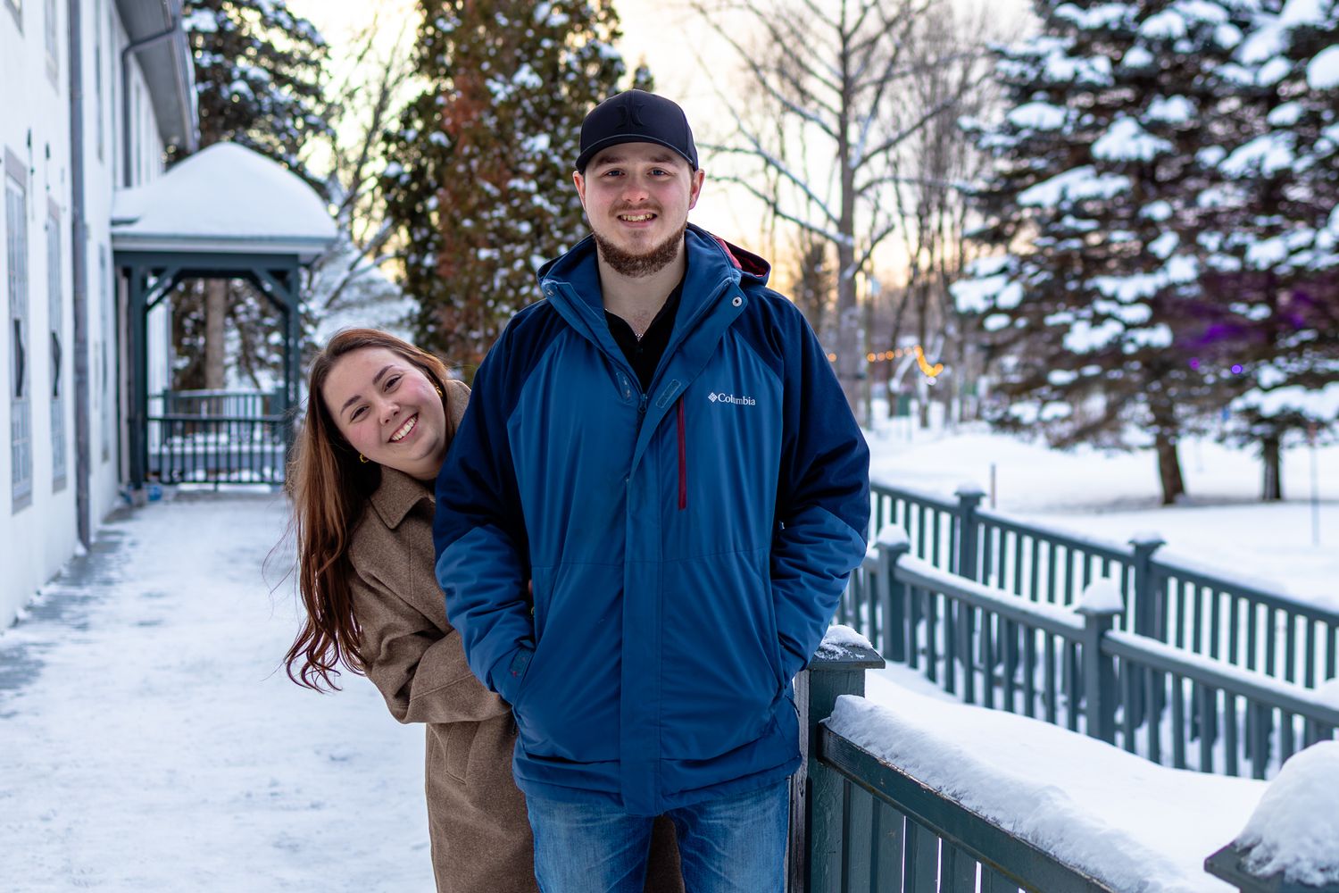 Happy couple smiling together on snowy winter day, woman in tan coat and man in blue Columbia jacket by white fence.