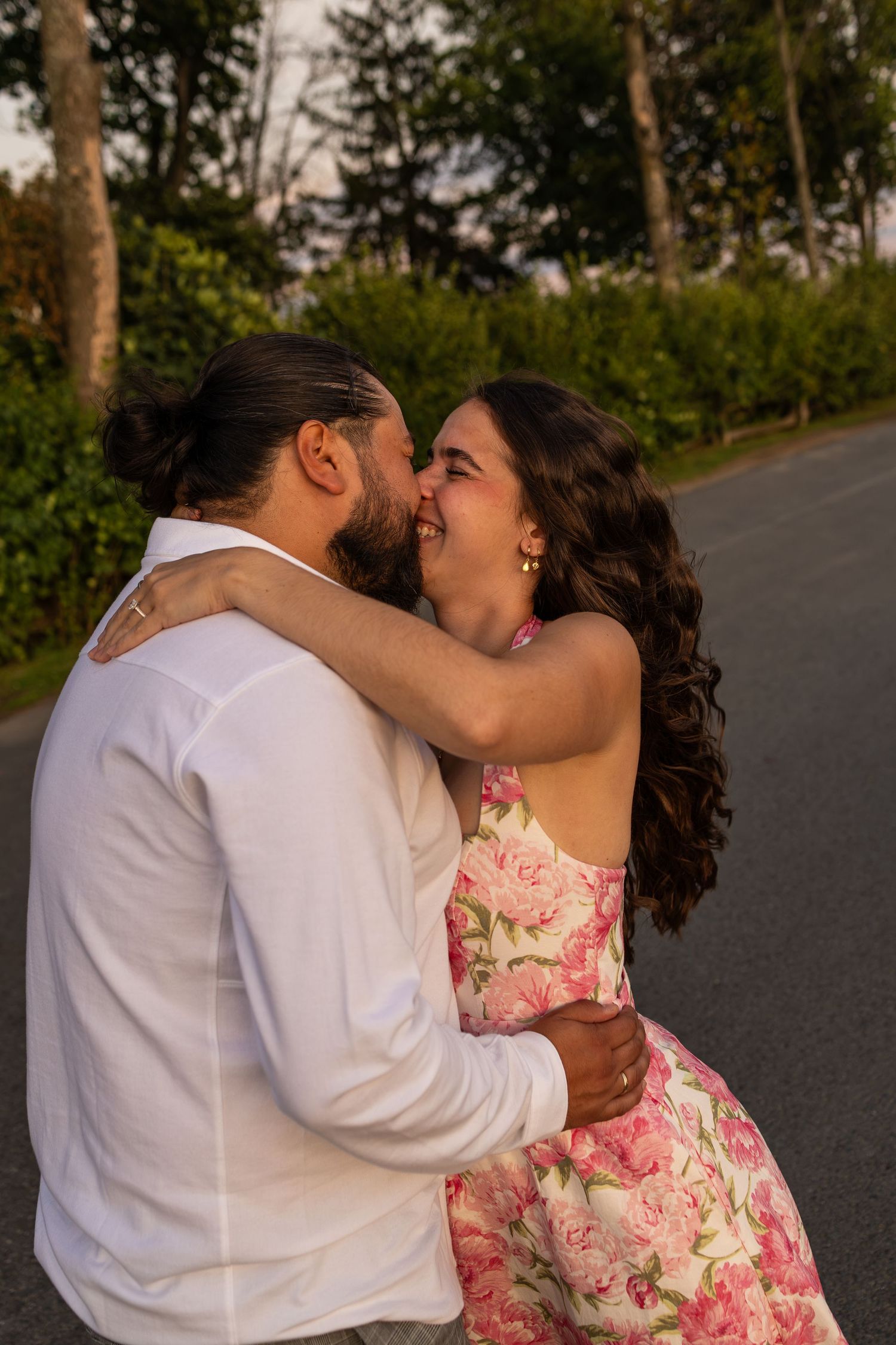 A couple shares a romantic embrace on a road, the woman in a floral pink dress laughing as the man kisses her cheek.
