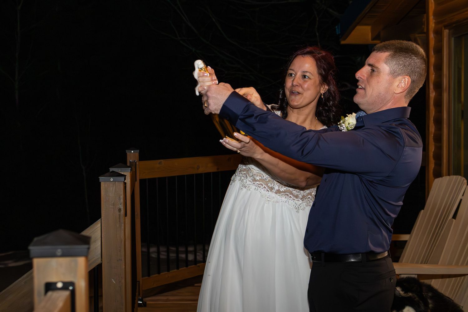 Newlywed couple celebrates outdoors at night by popping champagne bottle on wooden deck after wedding.