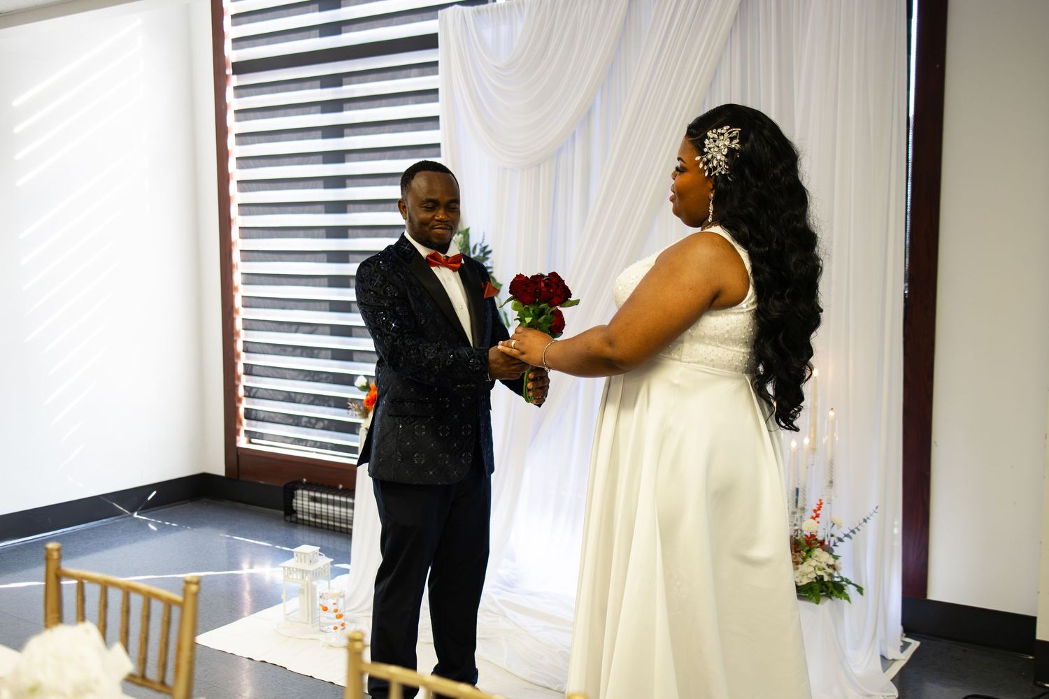 Bride and groom exchange vows at elegant indoor wedding ceremony with white drape backdrop and floral decorations.