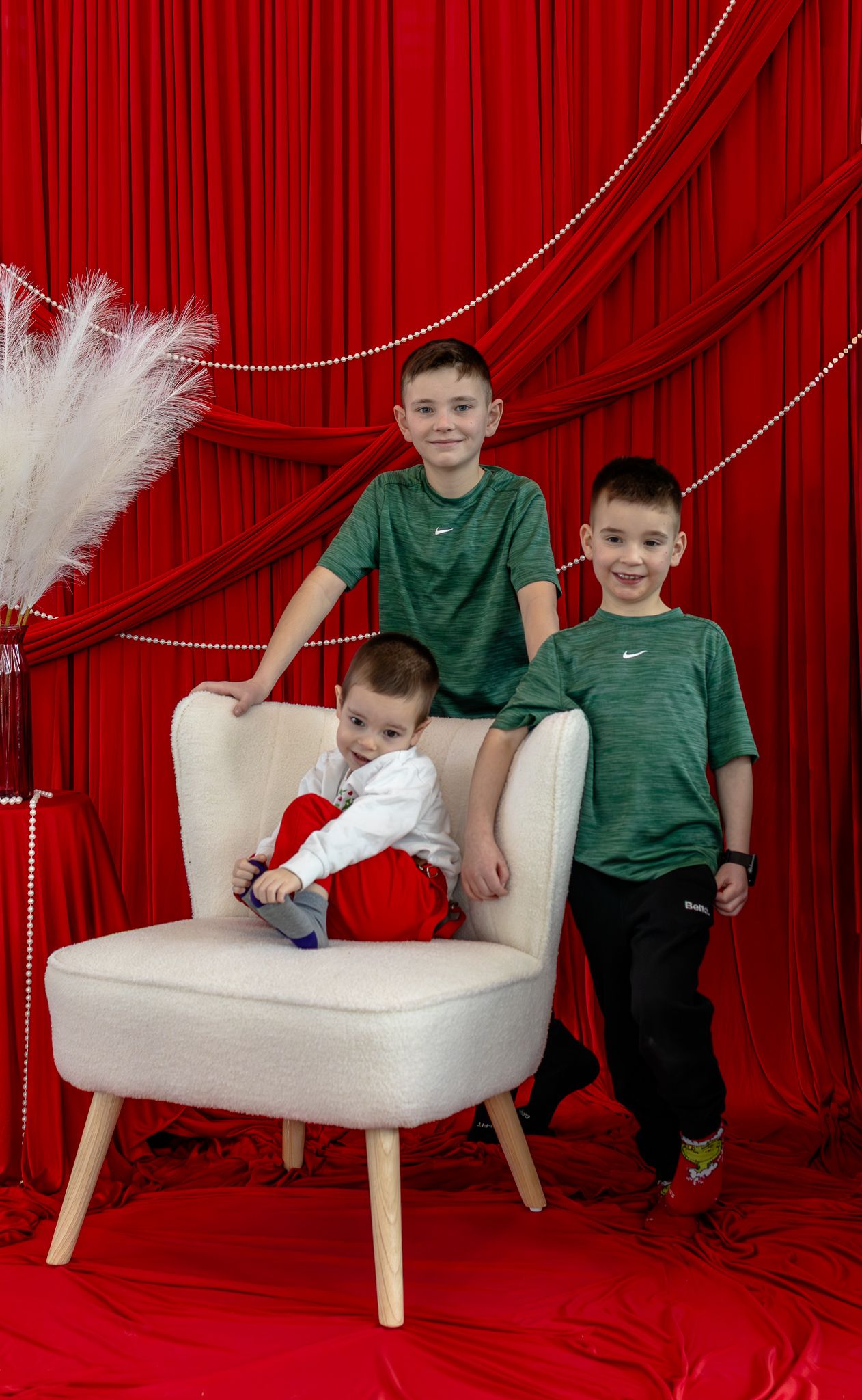 Three young brothers posing together with white armchair against festive red curtain background.