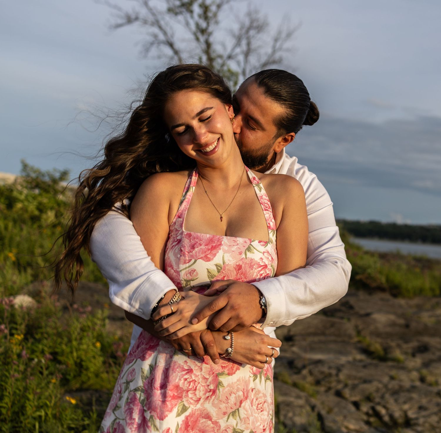 Couple embracing outdoors, woman in floral dress smiling, man in white shirt, rocky shoreline background.
