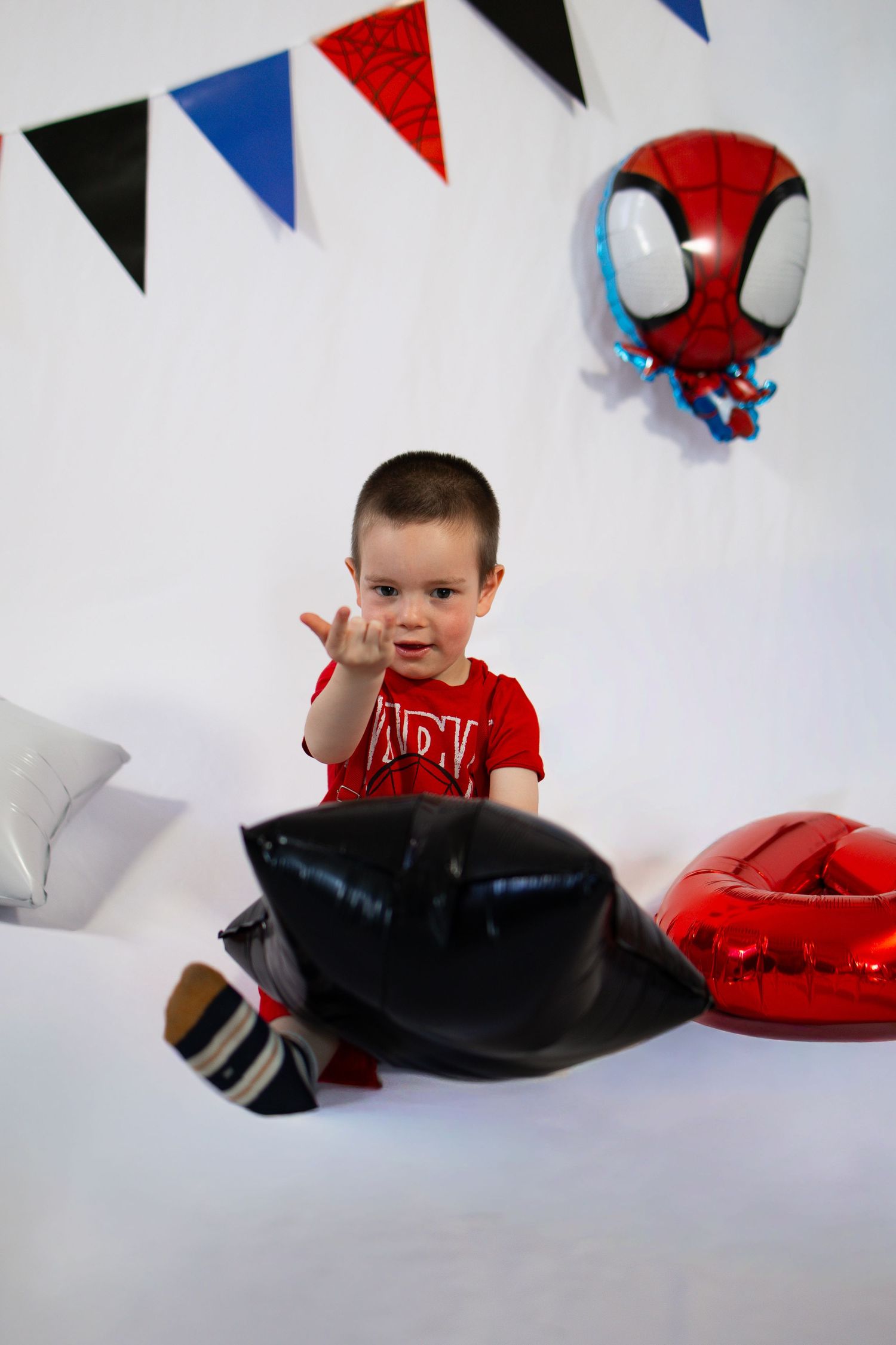 Young boy in red shirt sits on bed with black and red balloons at Spiderman themed birthday party.