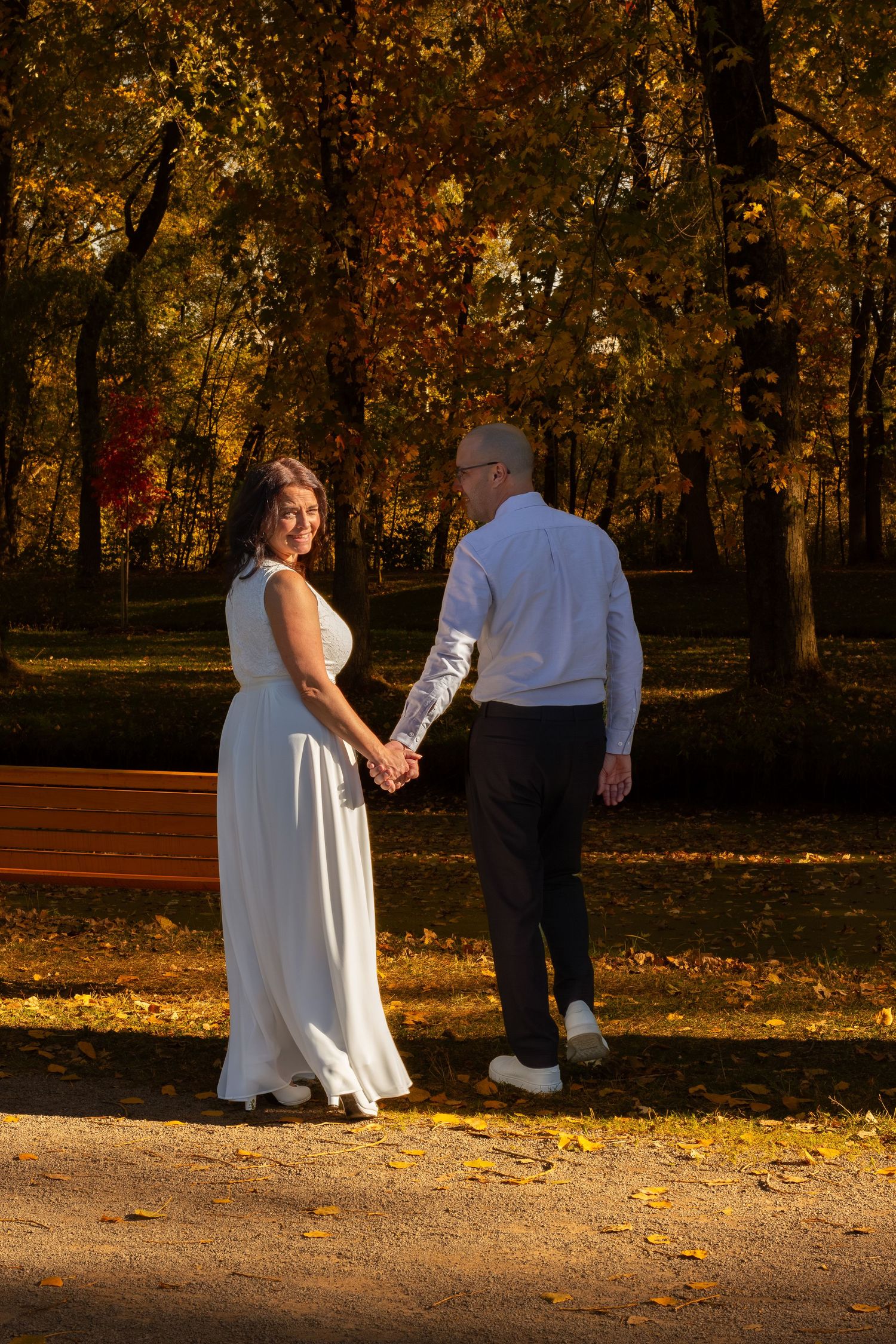 Bride in white dress and groom holding hands while walking through a sunlit autumn park.