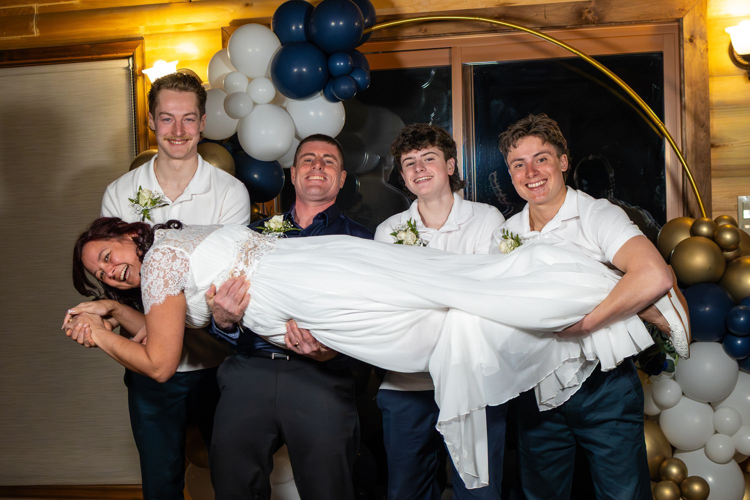 Wedding party playfully carrying bride in white dress in front of navy blue and gold balloon arch.