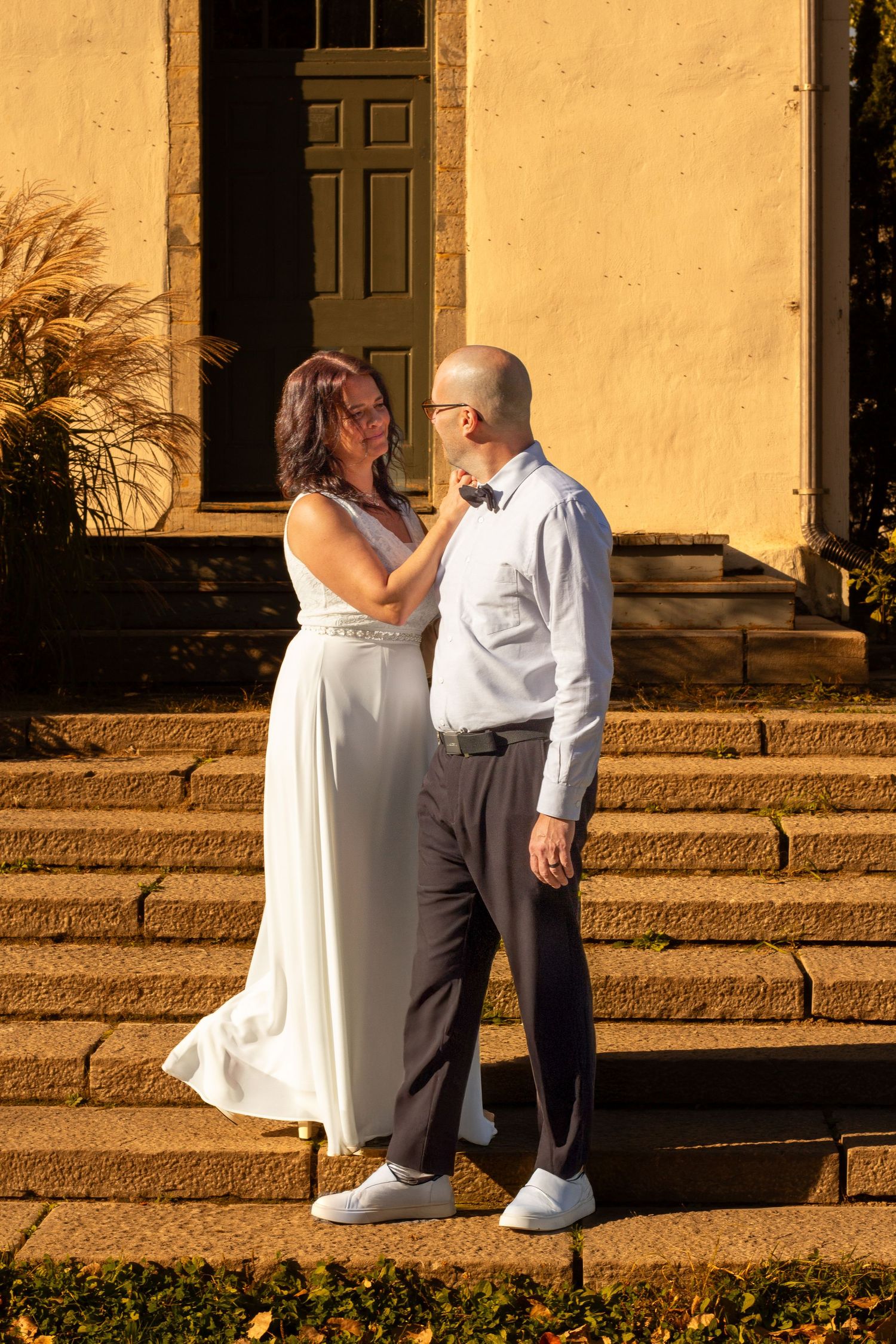 Couple in wedding attire standing on stone steps outside a yellow building in warm autumn sunlight.