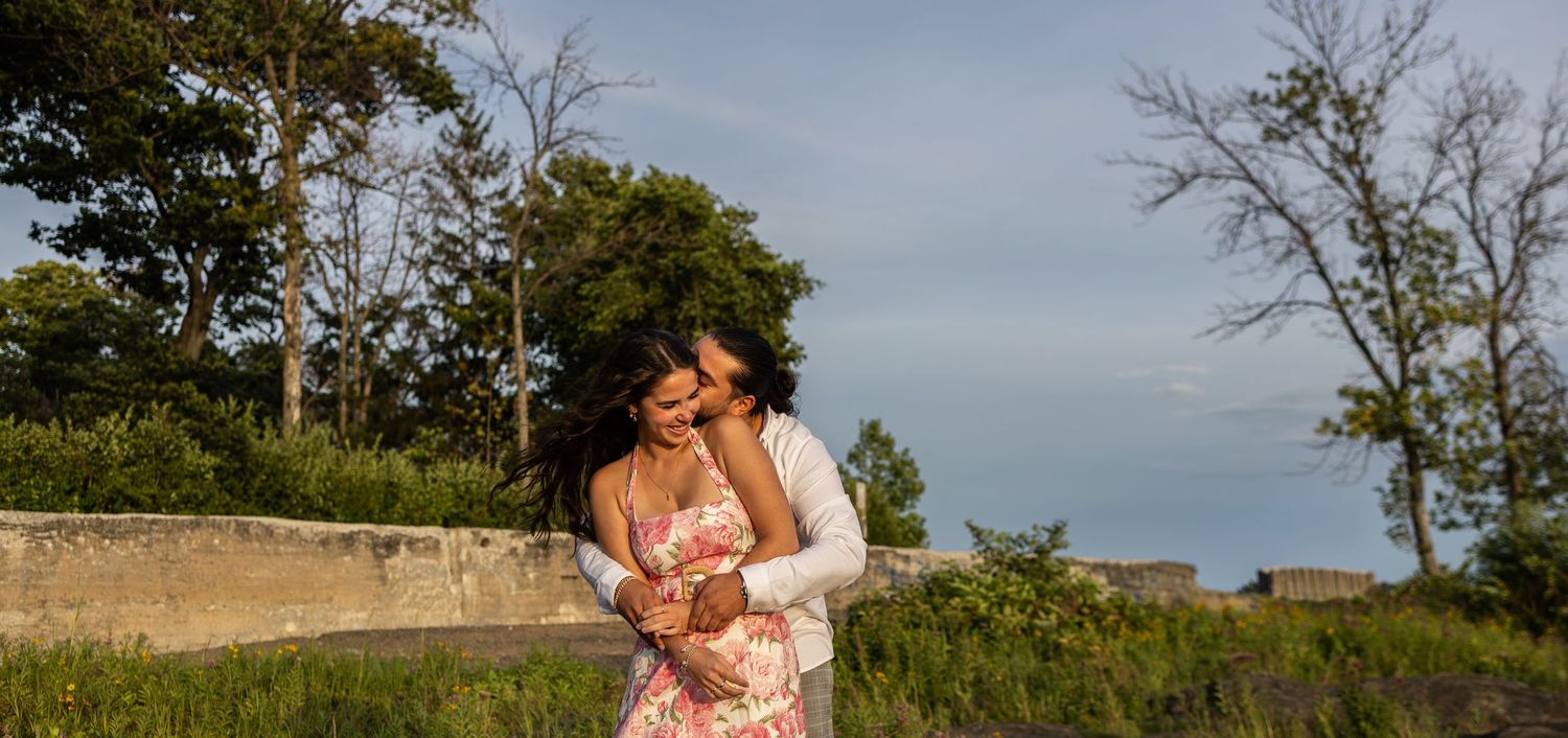 Couple embracing outdoors in a lush green field, woman in floral dress, man in white shirt, under a blue sky.