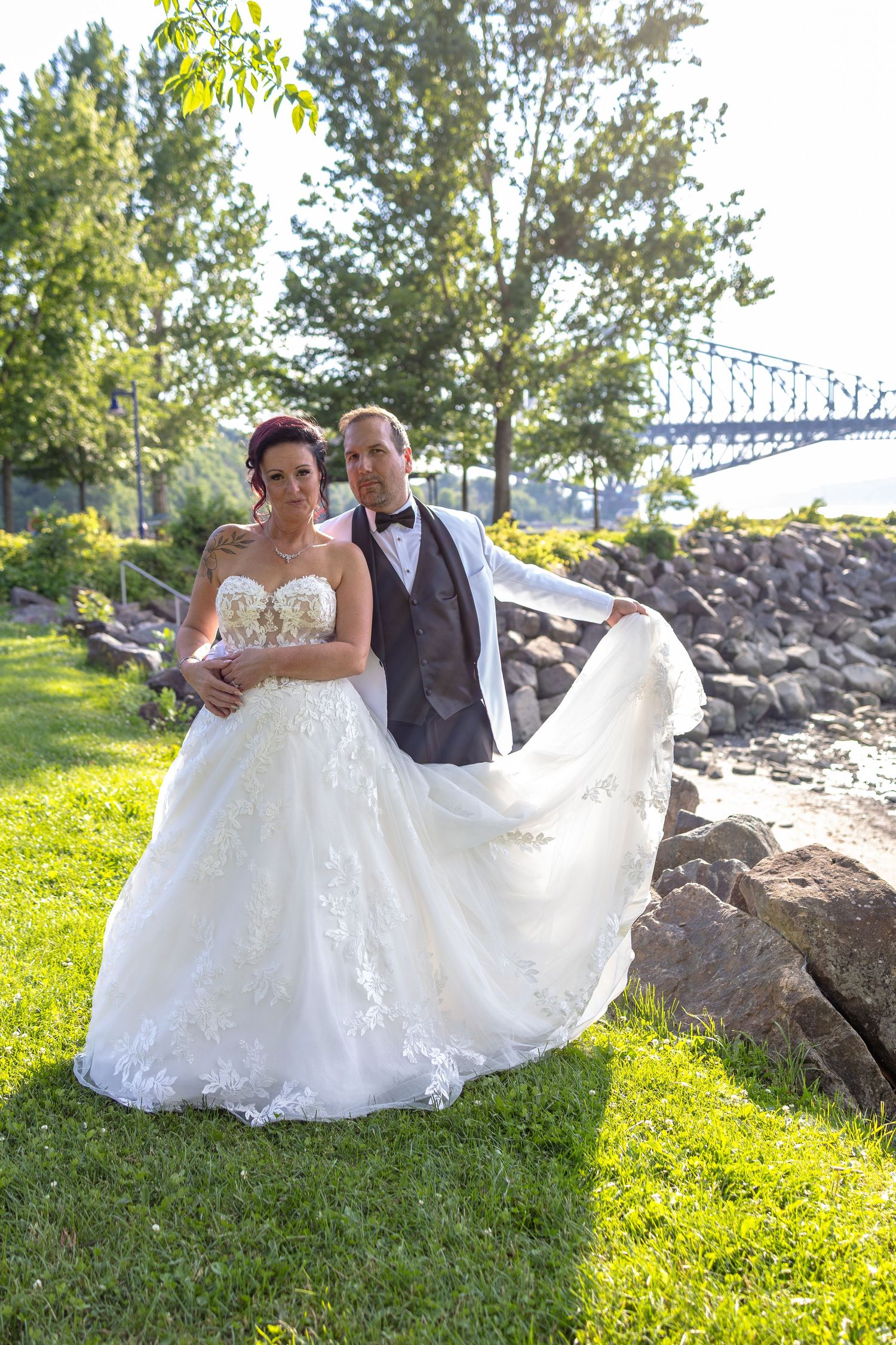 Bride and groom pose outdoors near rocky waterfront with bridge backdrop, groom lifting flowing white lace wedding gown train.