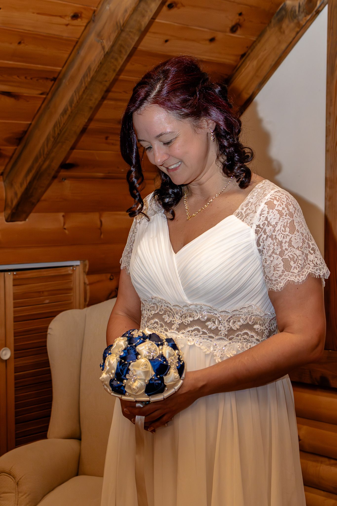 Bride in white lace wedding dress smiling while holding a navy blue and ivory floral bouquet in a log cabin.