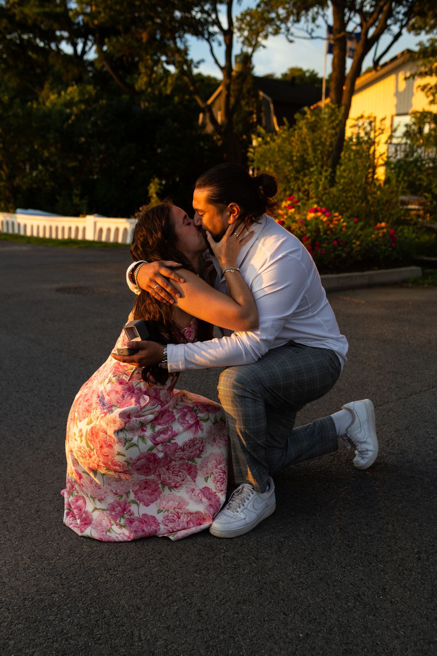 Couple sharing a romantic kiss outdoors at sunset, woman in floral dress, man in white shirt kneeling on pavement.